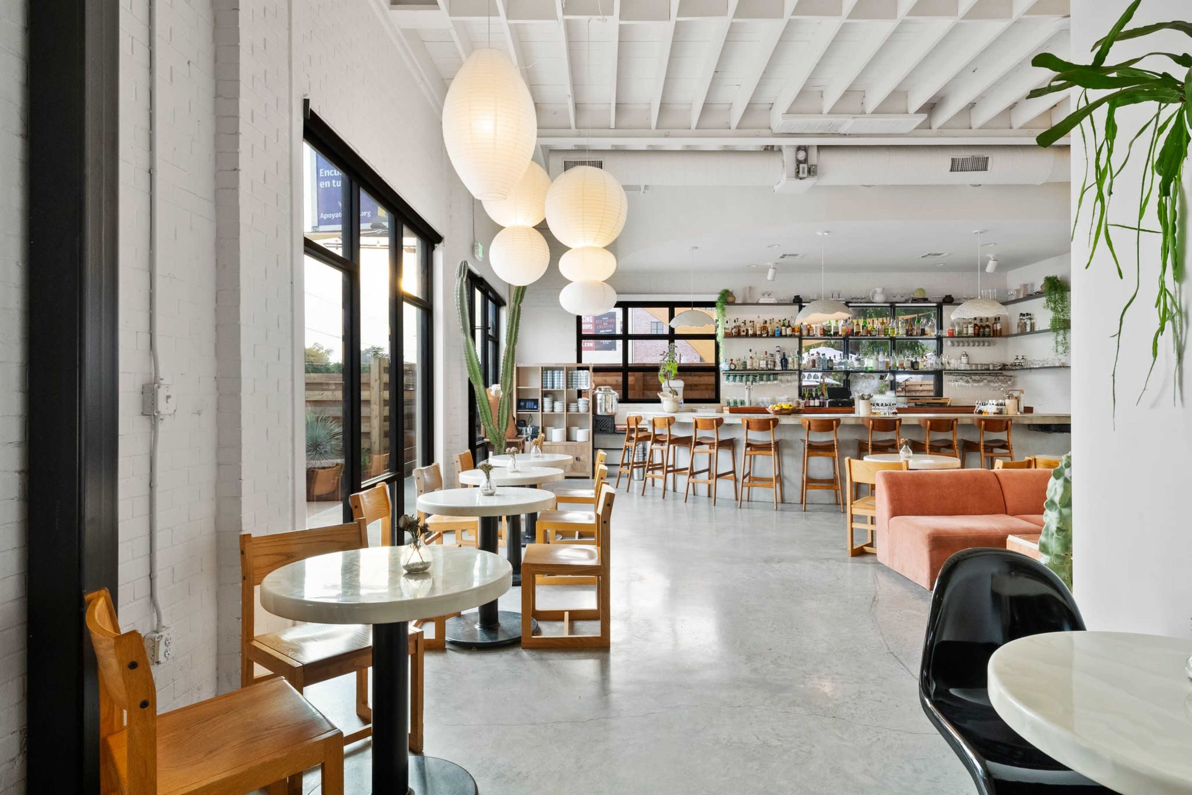 The image shows a modern restaurant interior with wooden tables, a bar area in the background, and large round light fixtures hanging from the ceiling.