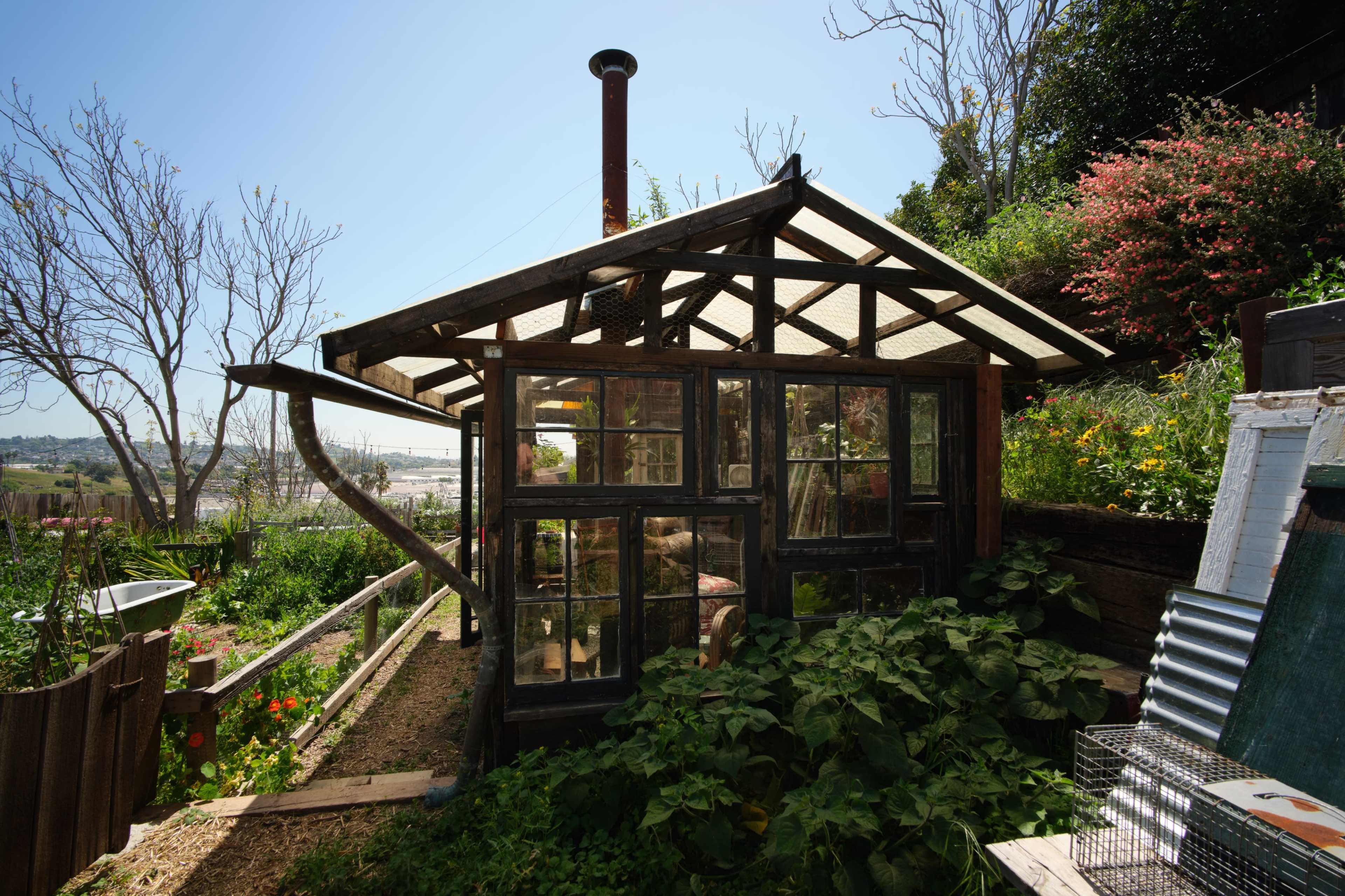 A small greenhouse with large glass panels and a metal chimney is surrounded by greenery and garden beds.