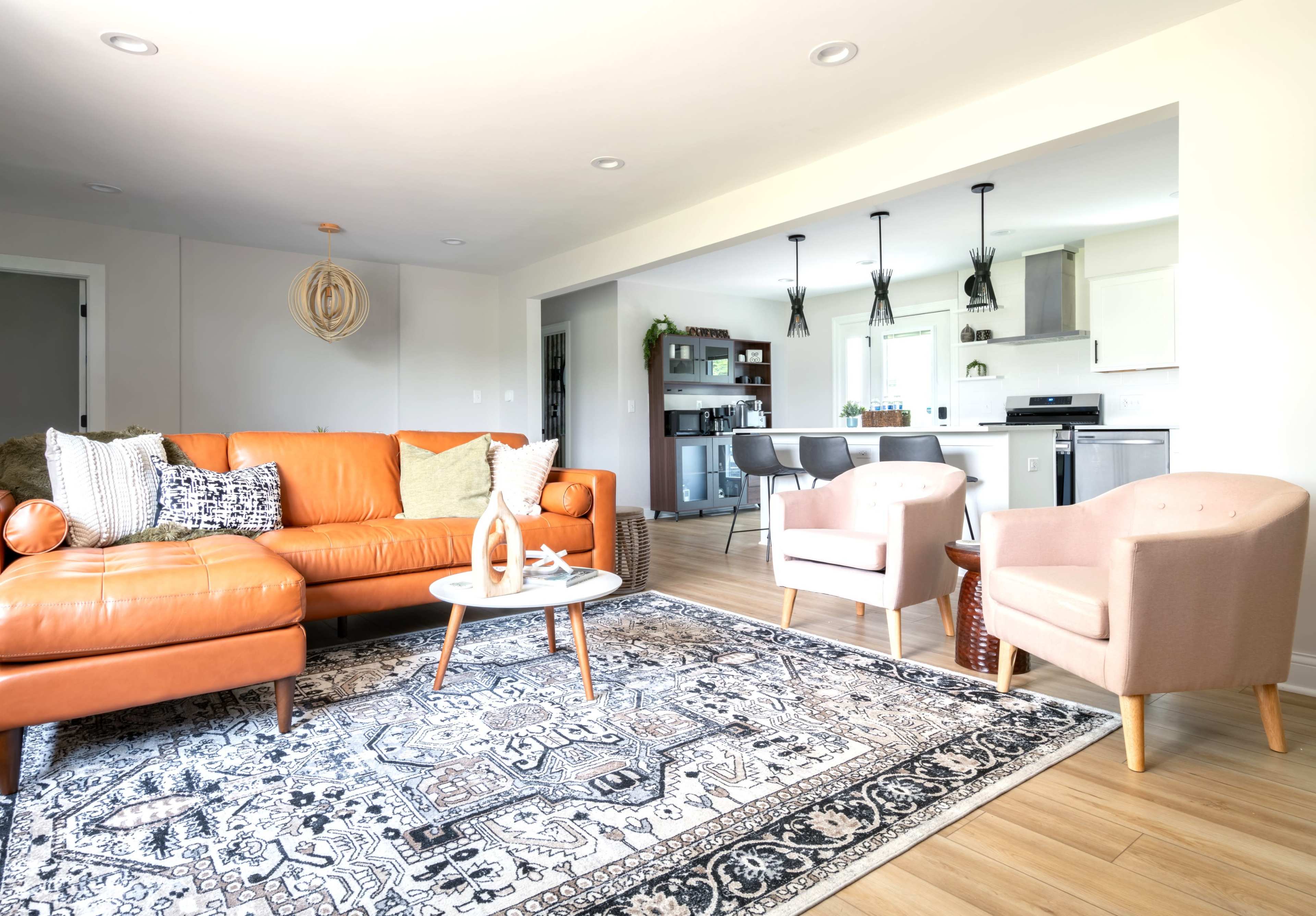 A modern living room features an orange leather sofa, two light pink armchairs, a decorative rug, and a view of a kitchen area in the background.