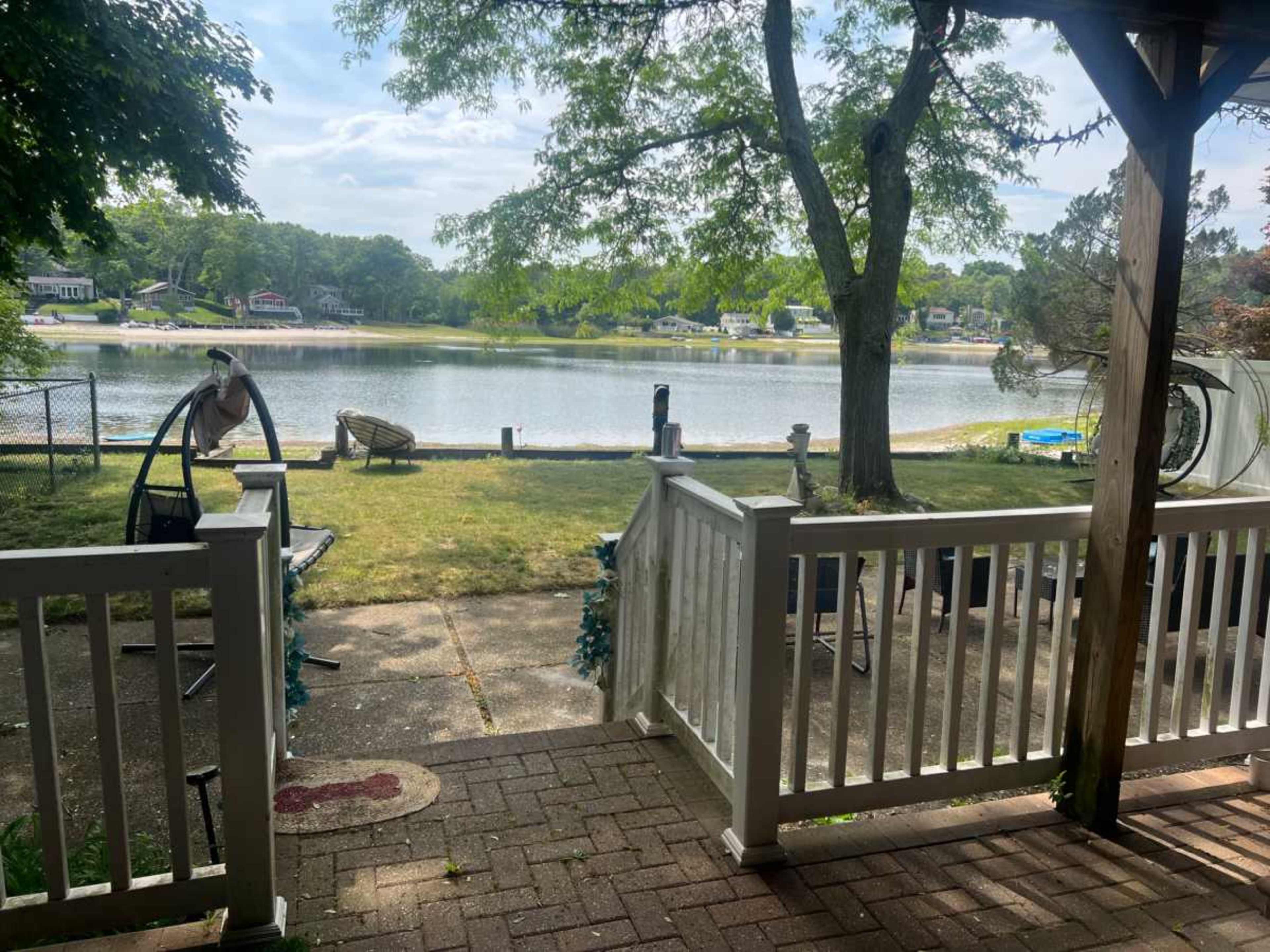 The image shows a view of a calm lake from a porch, with a grassy area and a few chairs visible in the foreground.