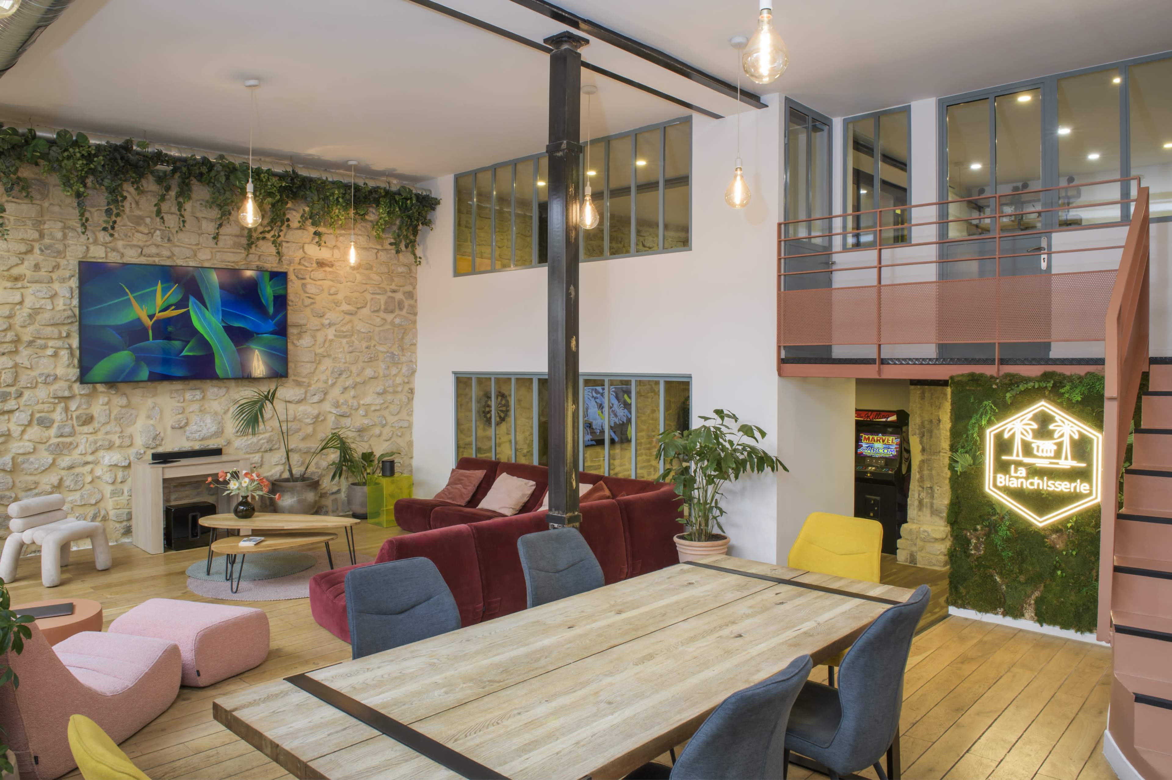 The image shows a modern communal living space featuring a wooden table, colorful seating areas, a large television, and plants decorating the walls.