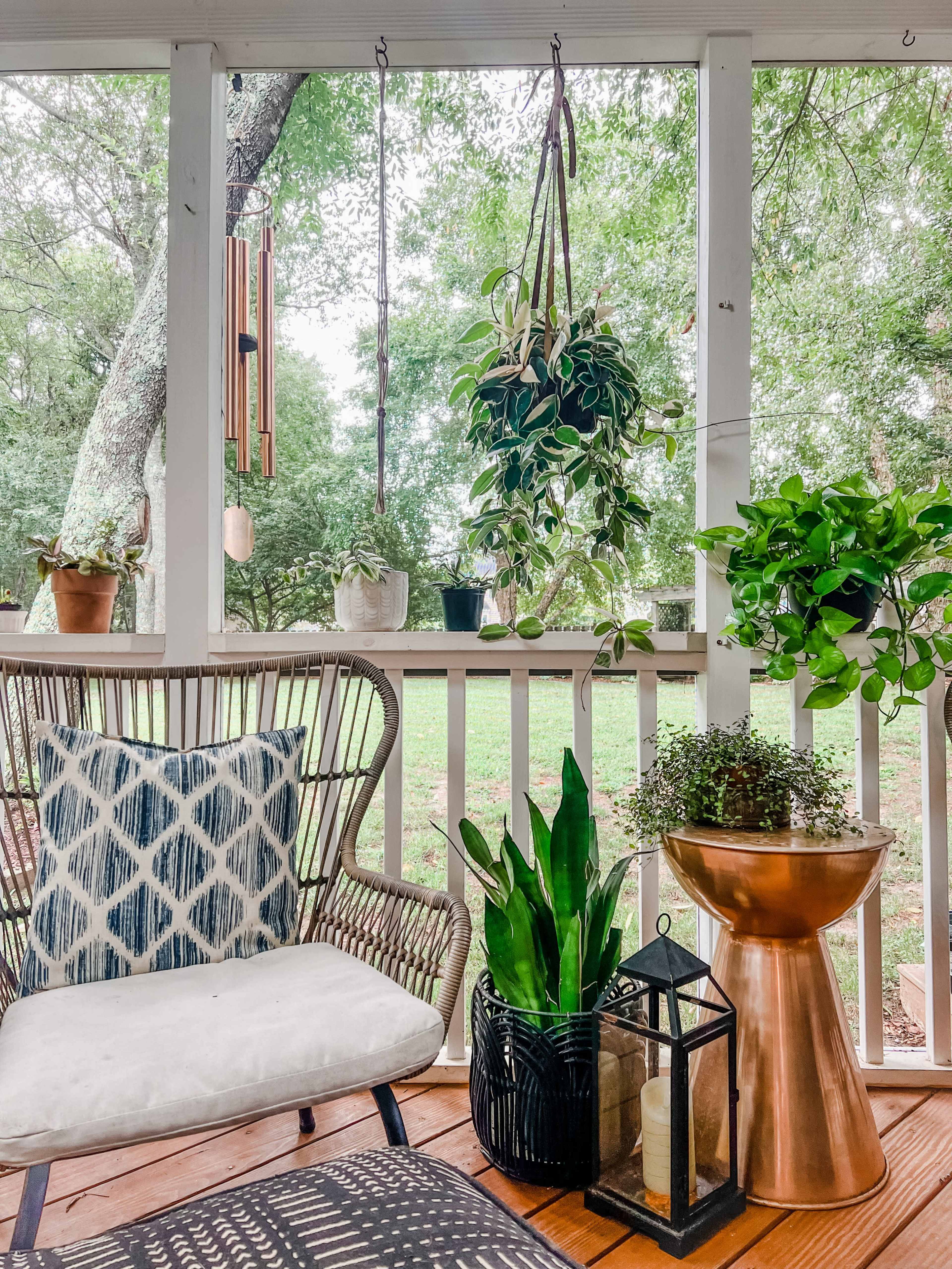 A cozy porch features a woven chair, hanging plants, potted greenery, and a decorative lantern on a wooden floor.