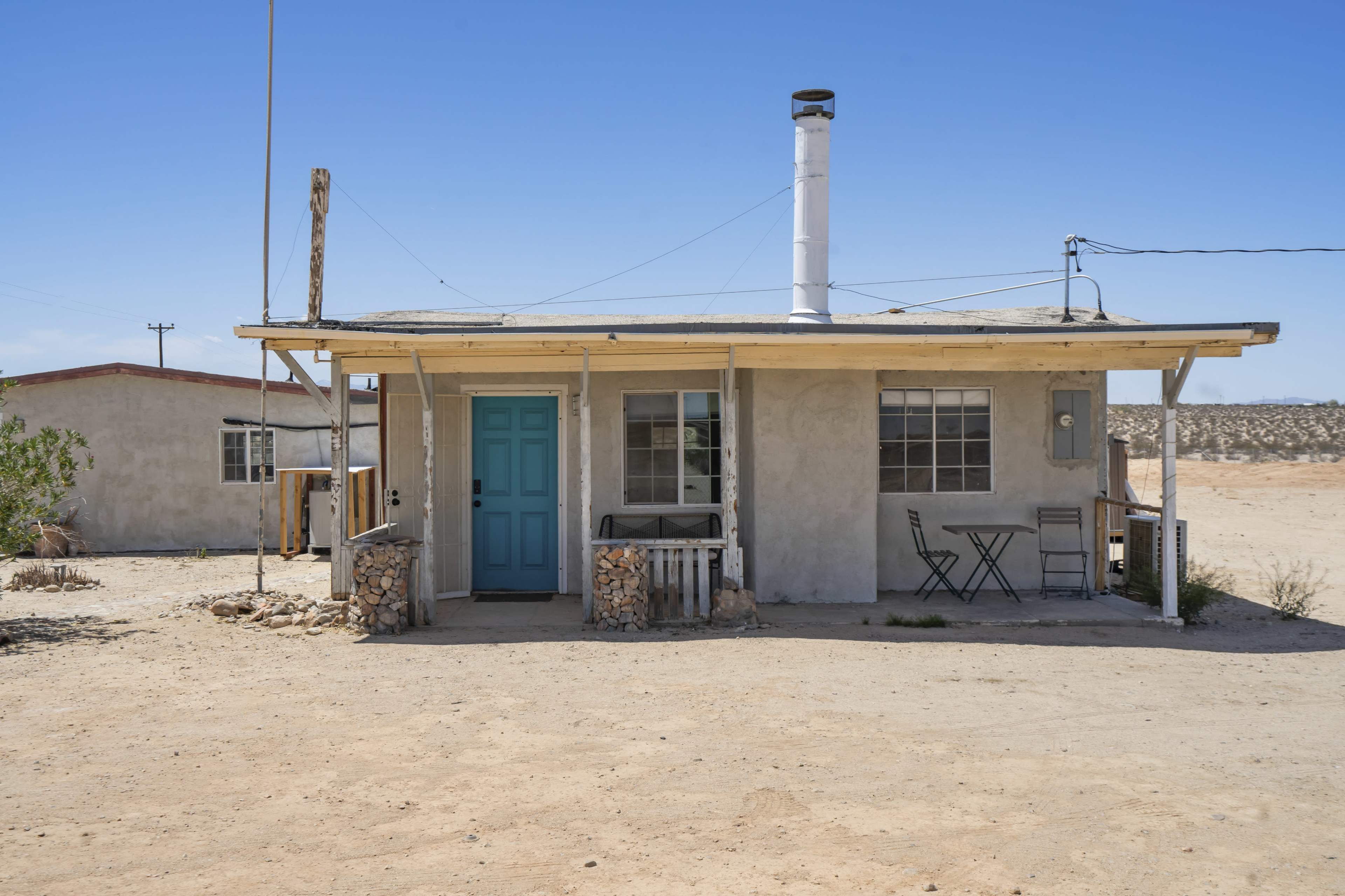 A small, light-colored house with a blue door and a porch, surrounded by a sandy landscape.