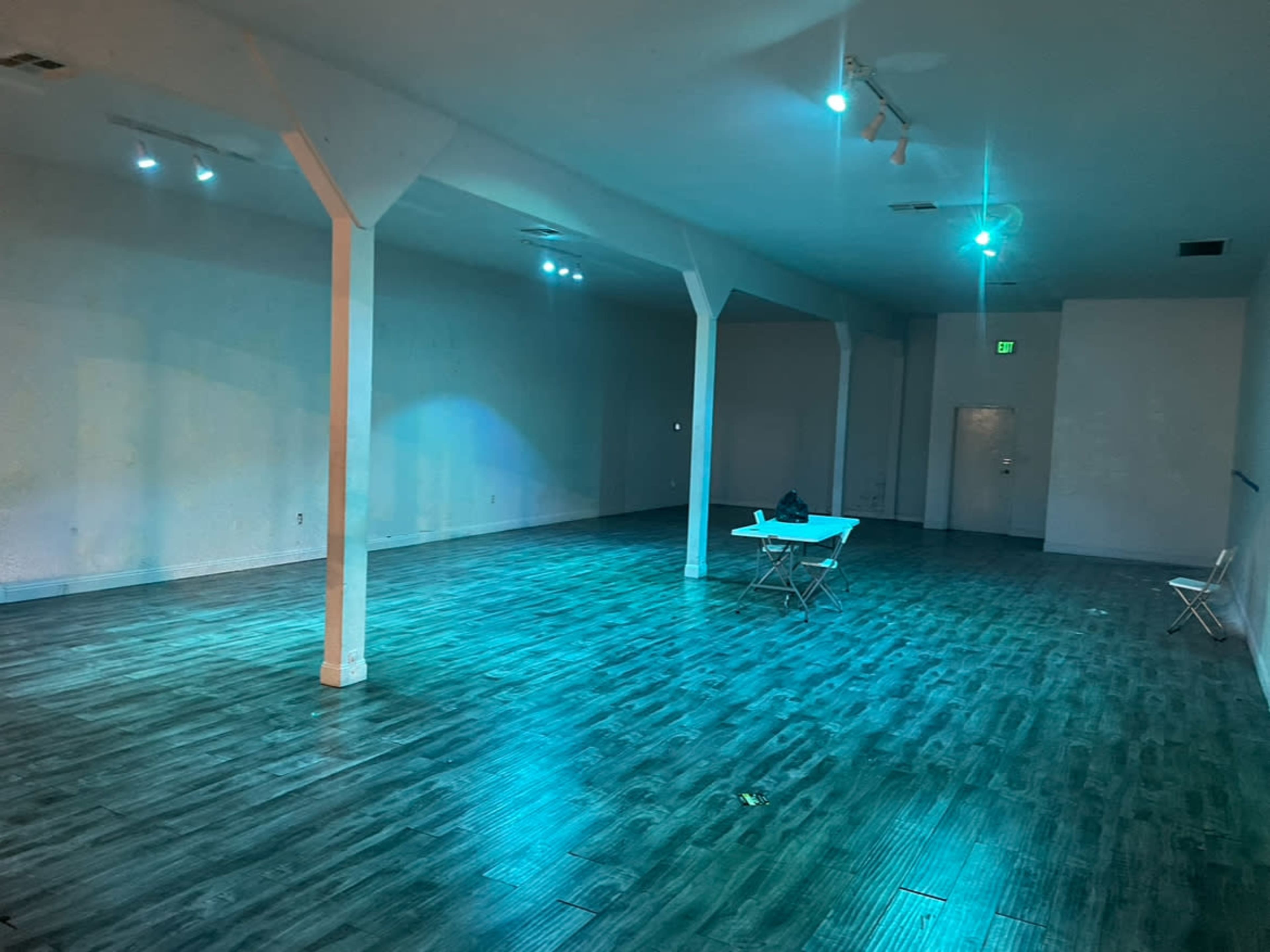 An empty room with a wooden floor, illuminated by soft blue lights, featuring a table and chairs in the center.