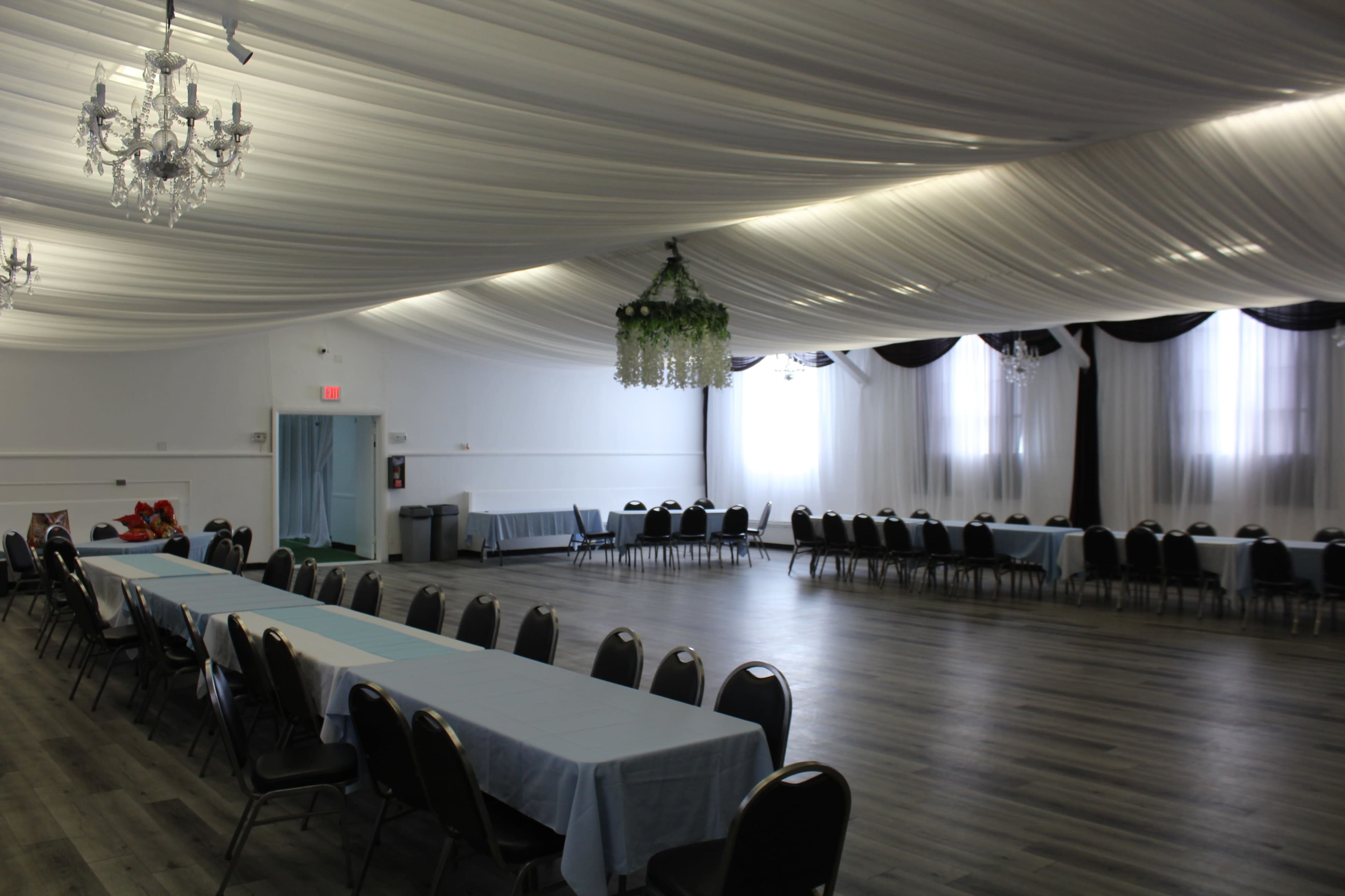 The image shows a spacious banquet hall with long tables covered in light blue fabric, surrounded by black chairs under a draped ceiling.