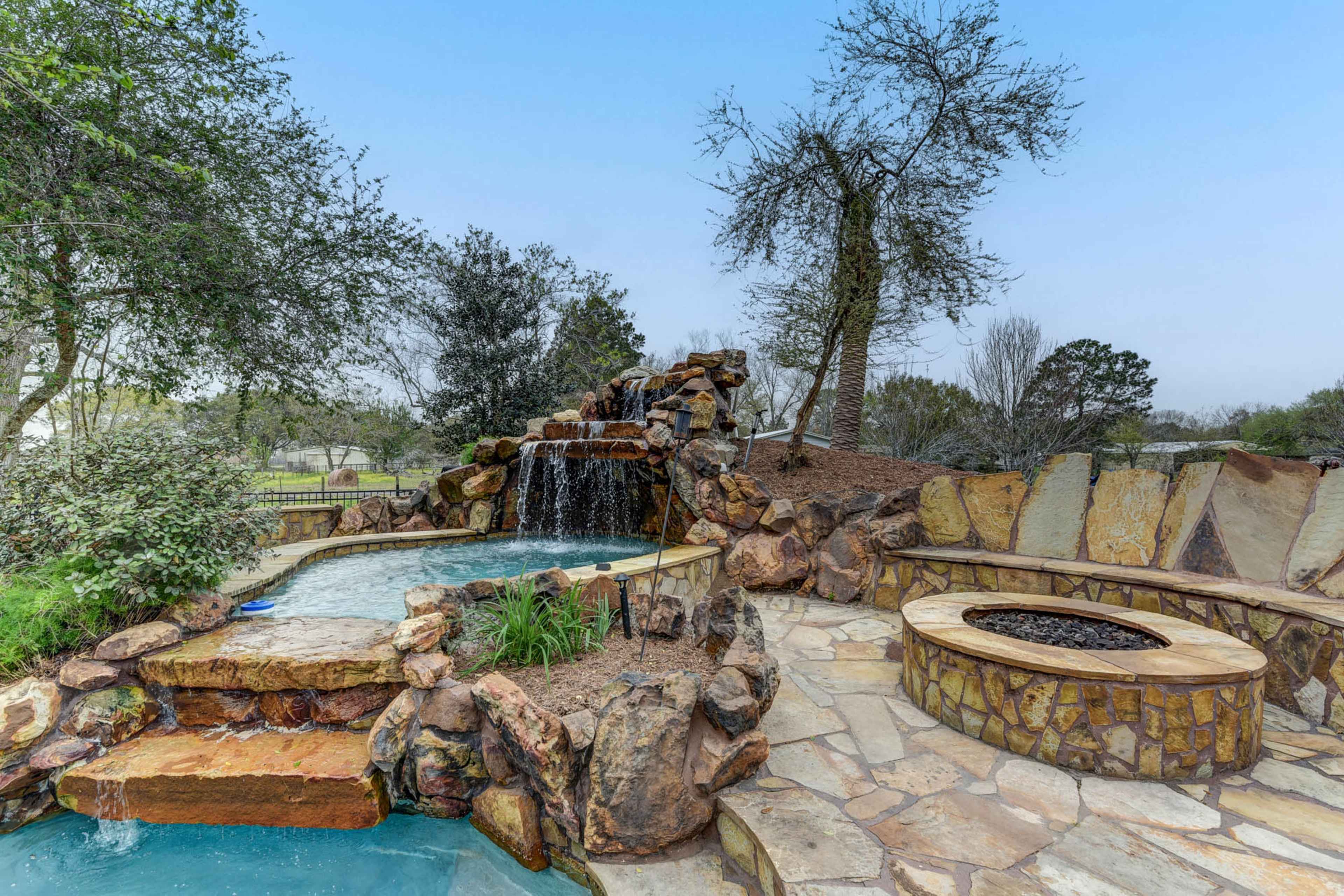 A stone waterfall cascading into a small pool, surrounded by landscaped greenery and a fire pit beside a stone walkway.
