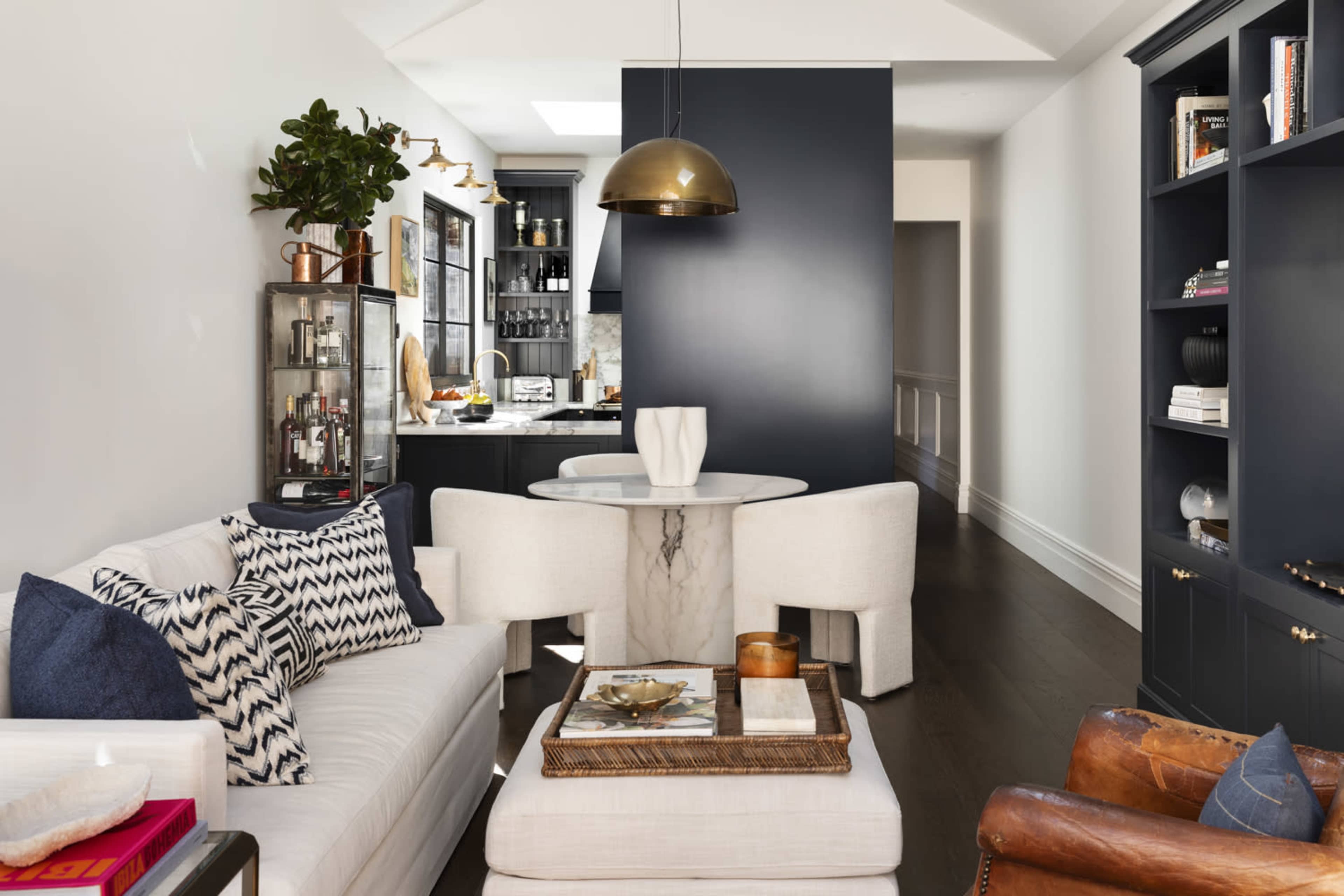 A modern living space features a white sofa with patterned cushions, a round marble table, and a dark accent wall leading to a kitchen area.