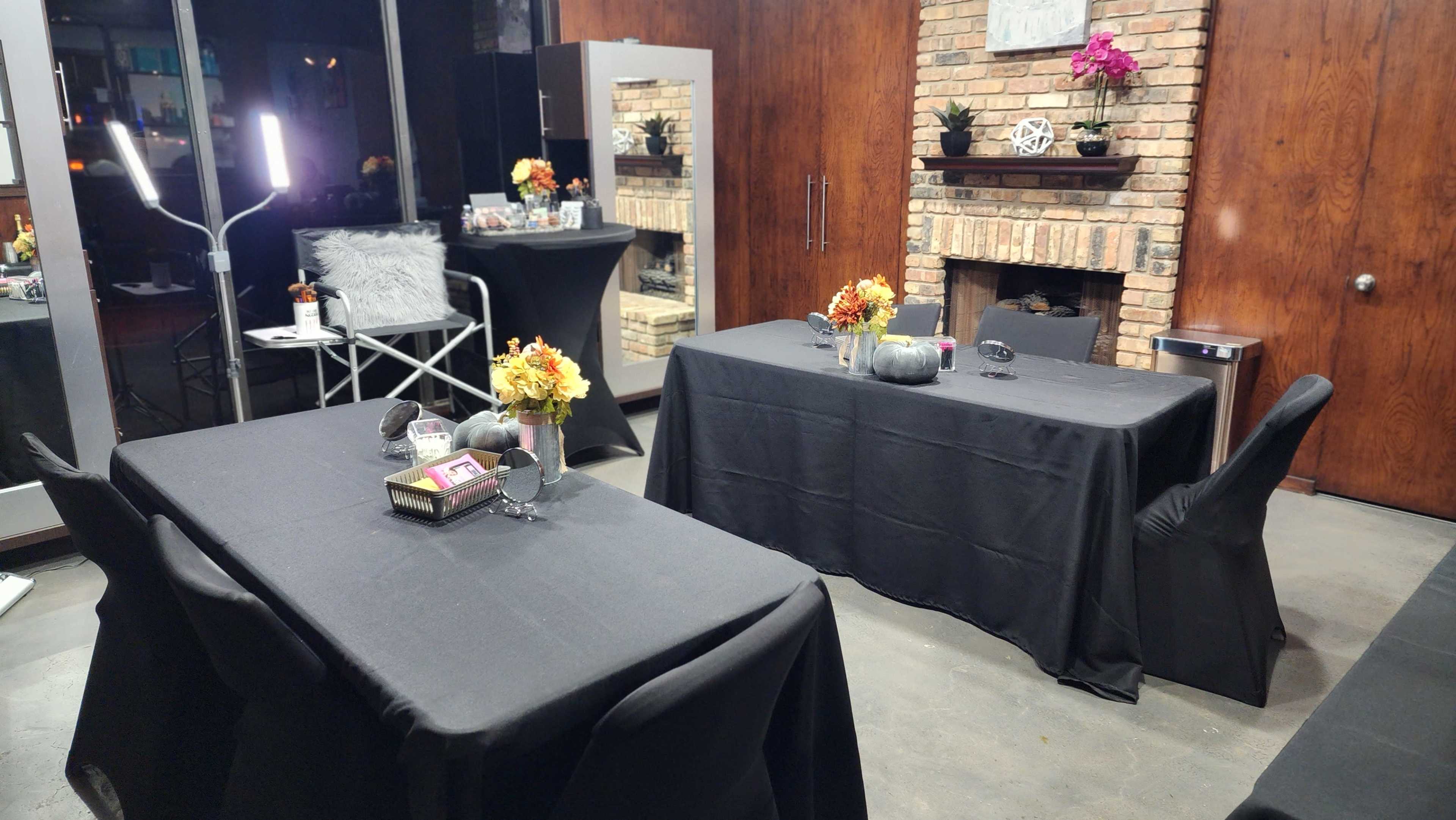 The scene features three tables with black tablecloths, adorned with floral arrangements, set up in a room with a brick fireplace and mirrored cabinets.