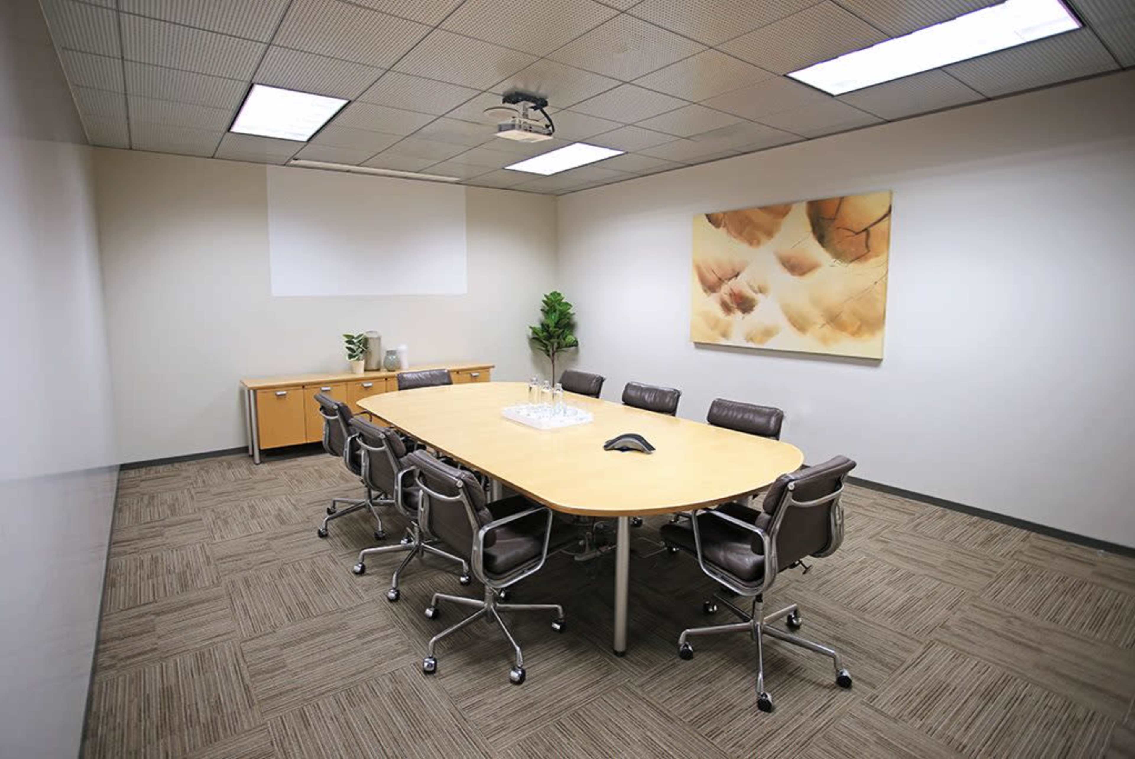 A modern conference room features a long oval table surrounded by ergonomic chairs, with a projector mounted on the ceiling and a large artwork on the wall.