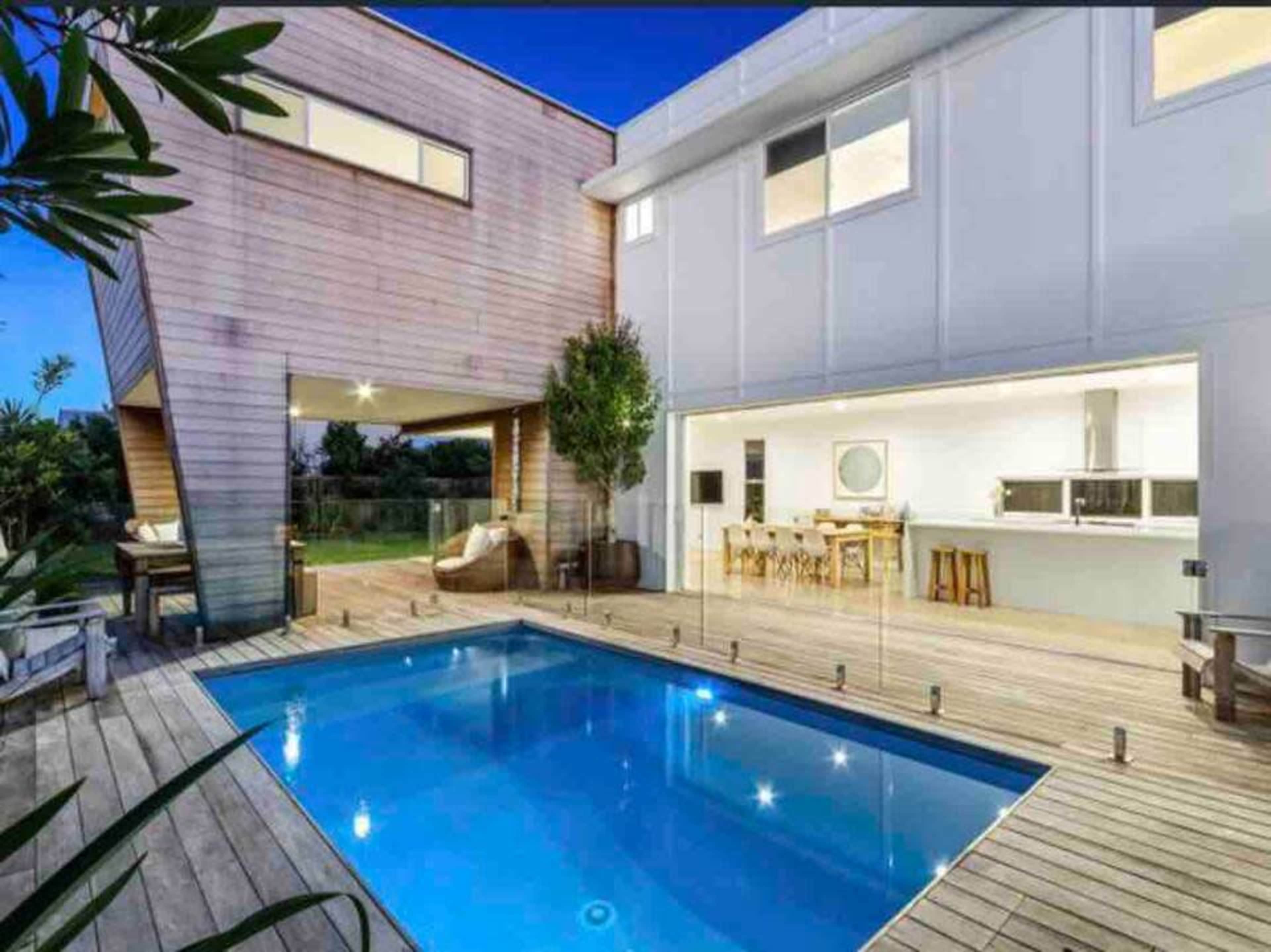 A modern house with a wooden deck and a rectangular swimming pool in the foreground, illuminated by lights against a twilight sky.