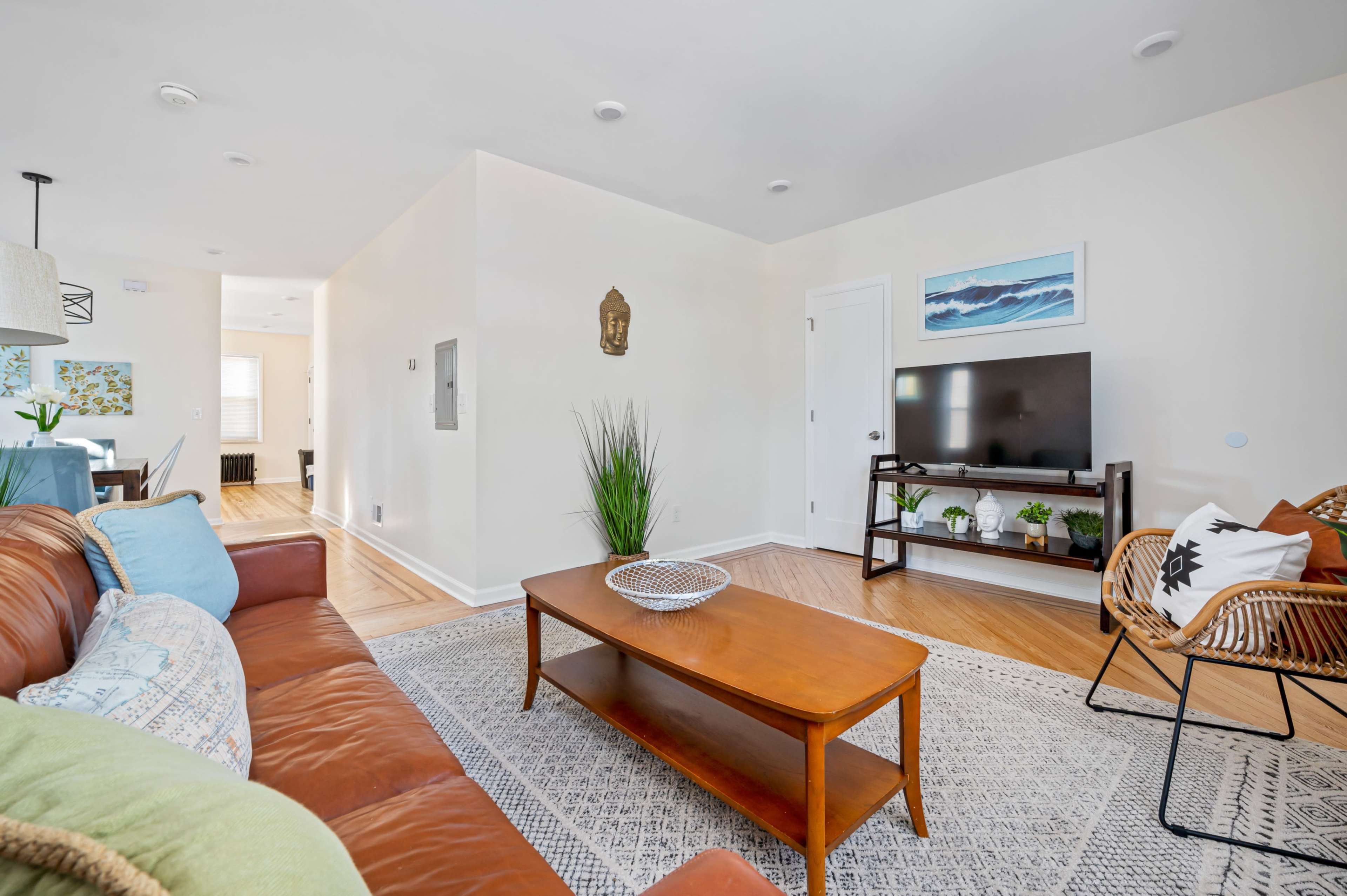 A modern living room with a brown leather sofa, a wooden coffee table, and a television on a stand, accented by decorative plants and artwork on the walls.