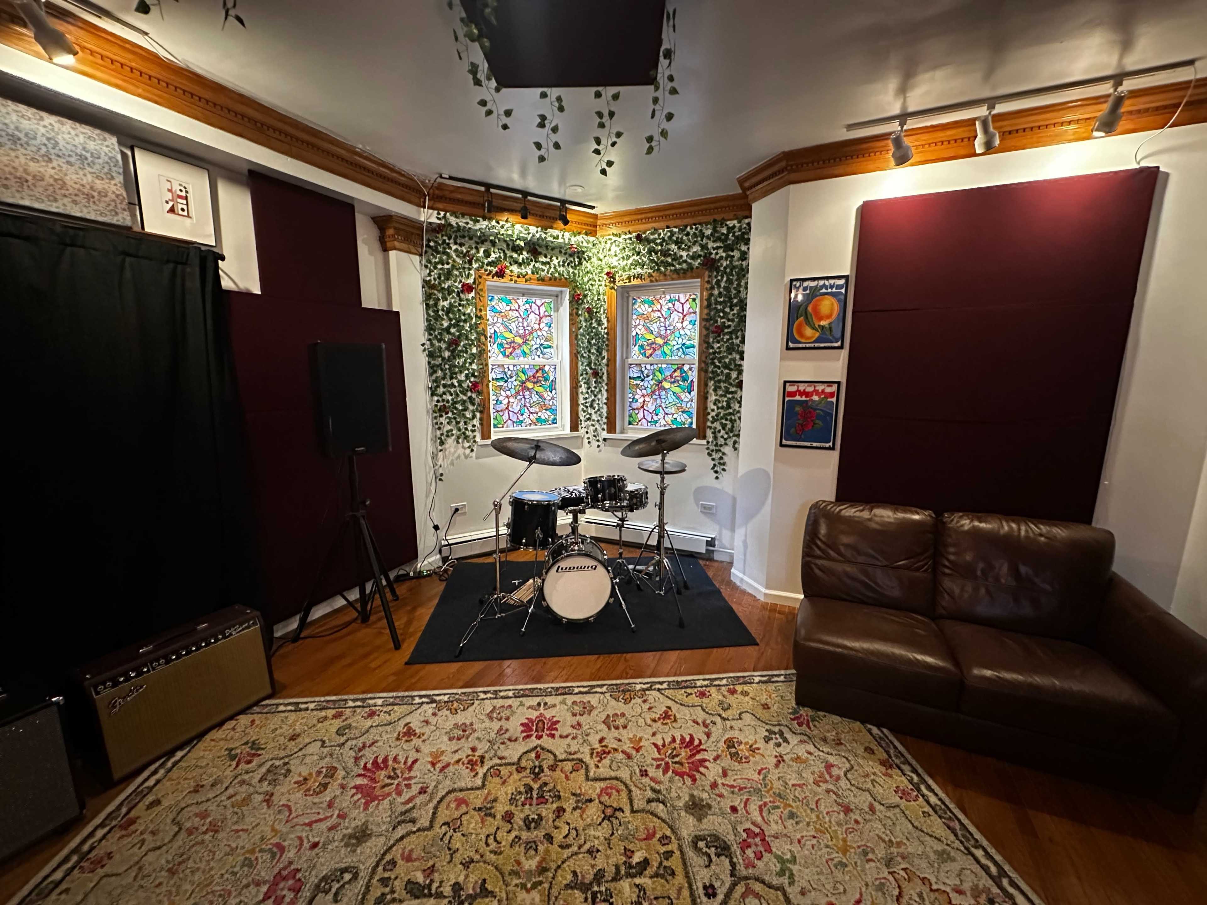 The image shows a music studio with a drum set positioned in front of two stained glass windows, surrounded by greenery and featuring a brown couch and amplifiers.