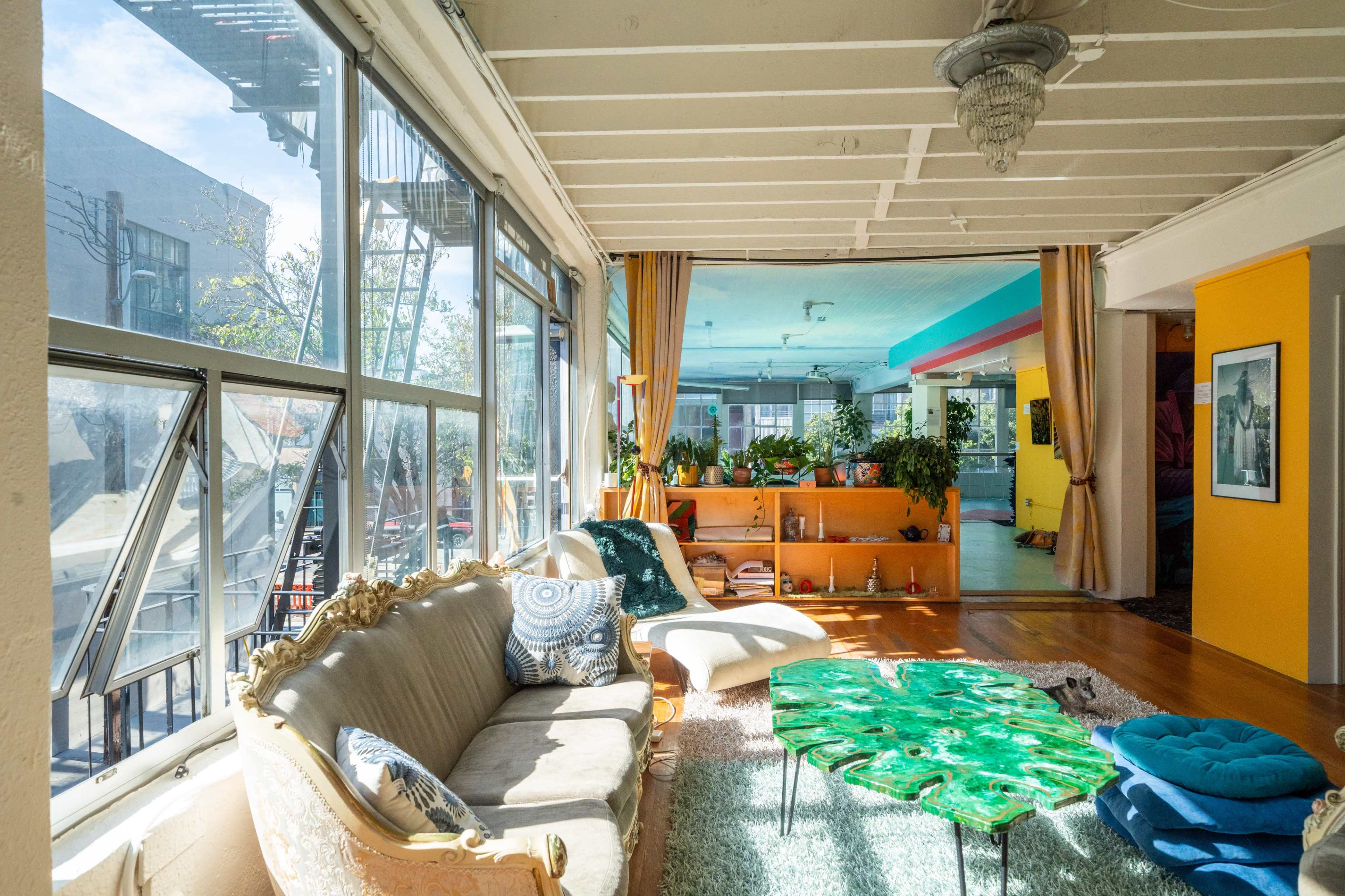 The image shows a bright, open living space with large windows, featuring a vintage sofa, a green decorative table, and various plants arranged throughout the room.
