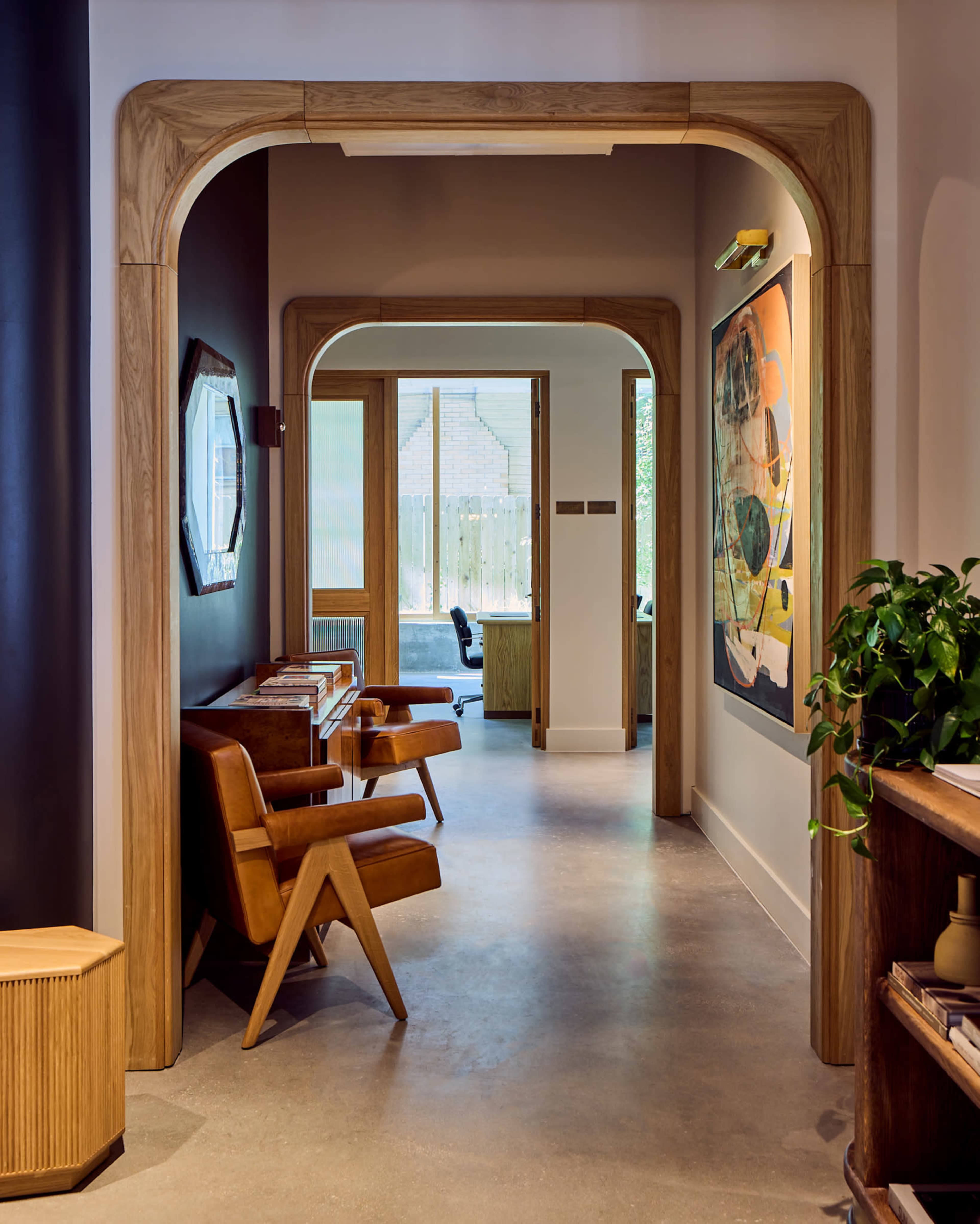 The image shows a modern hallway with an arched wooden doorway, featuring two stylish armchairs and a view into a well-lit room with large windows.
