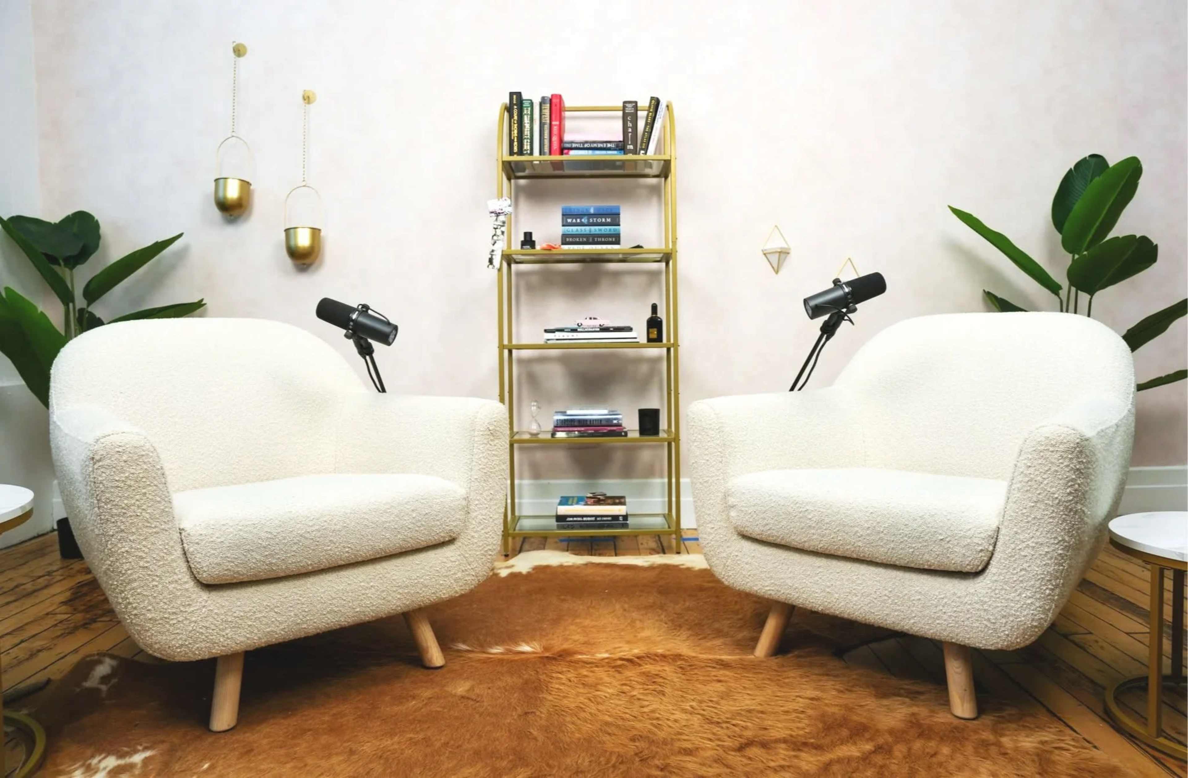 The image shows two white, upholstered chairs facing each other, placed on a cowhide rug, with a gold shelving unit in the background holding books and decorative items.