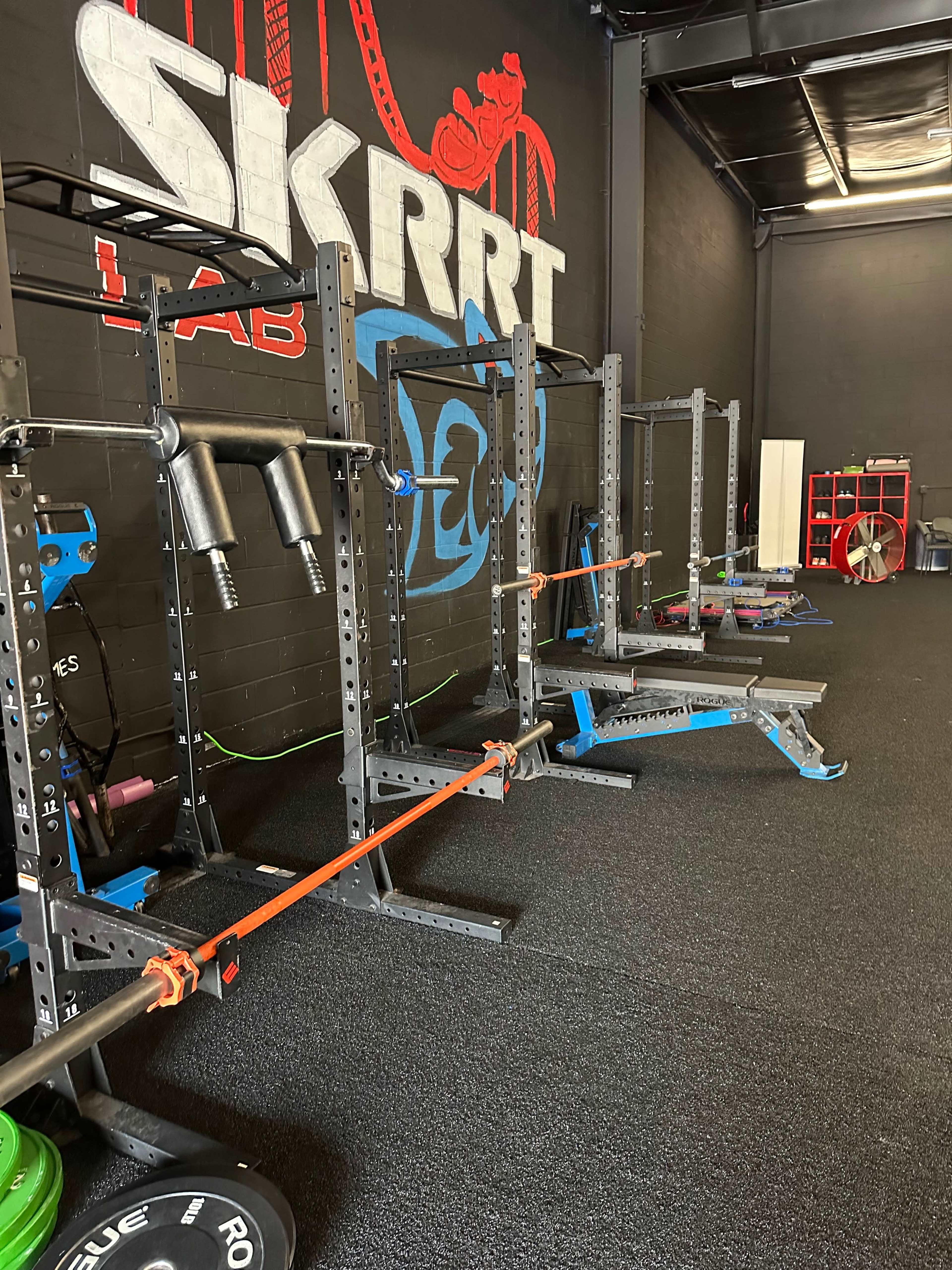 A gym interior featuring weightlifting equipment, including squat racks and a bench, against a black wall with the name "SKRRT LAB" painted in large letters.