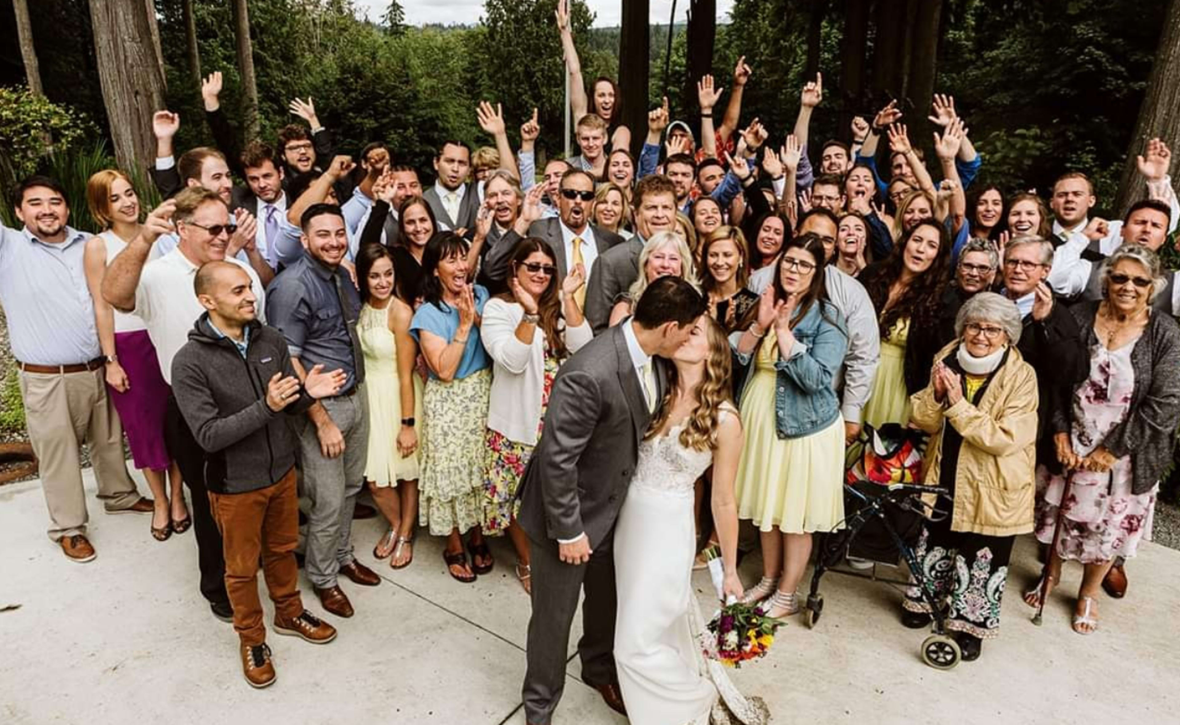 A large group of wedding guests poses for a photo outdoors, with the bride and groom at the center sharing a kiss.