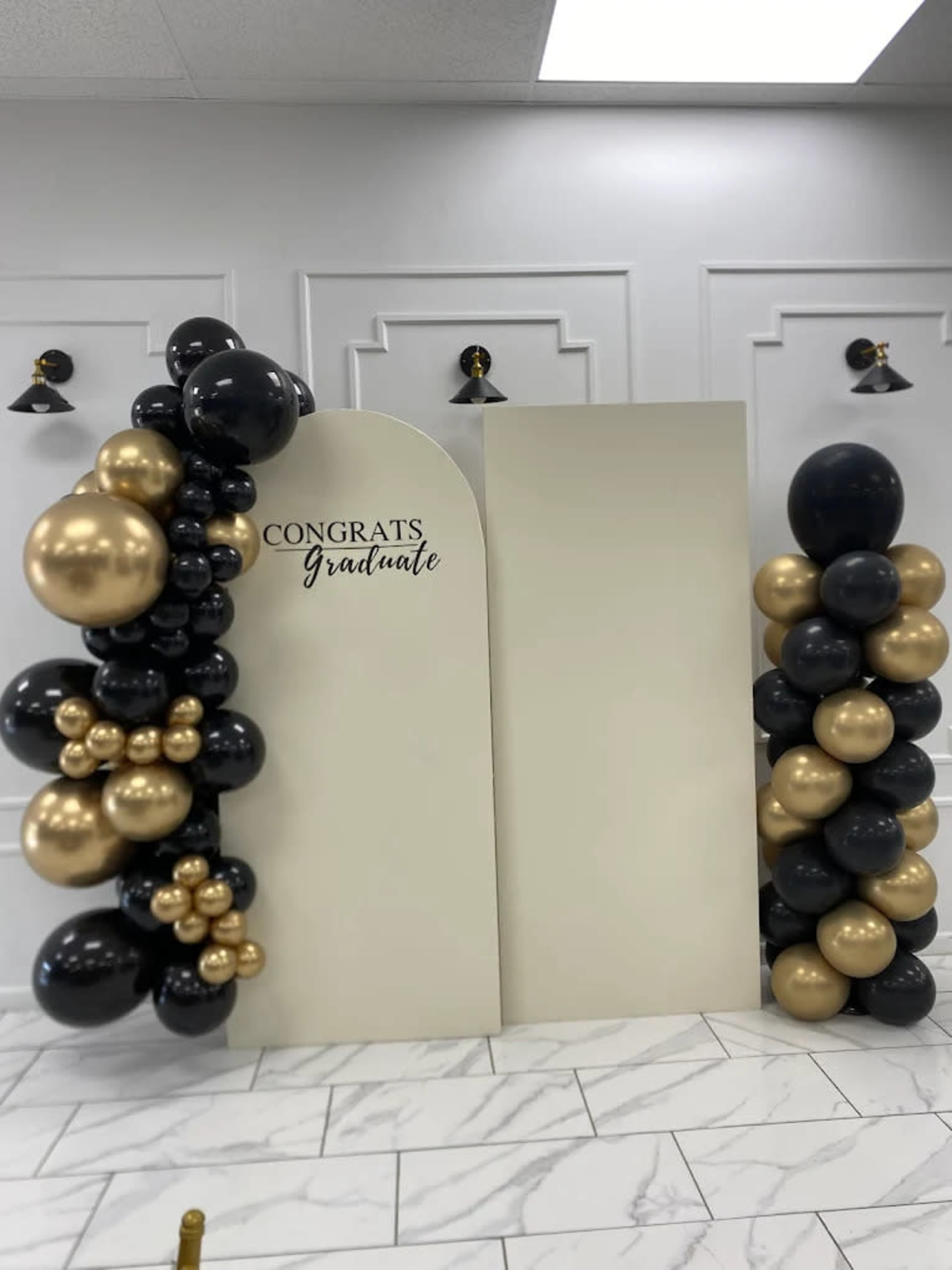 The image shows a decorated backdrop for a graduation celebration, featuring clusters of black and gold balloons alongside a sign that reads "CONGRATS Graduate."