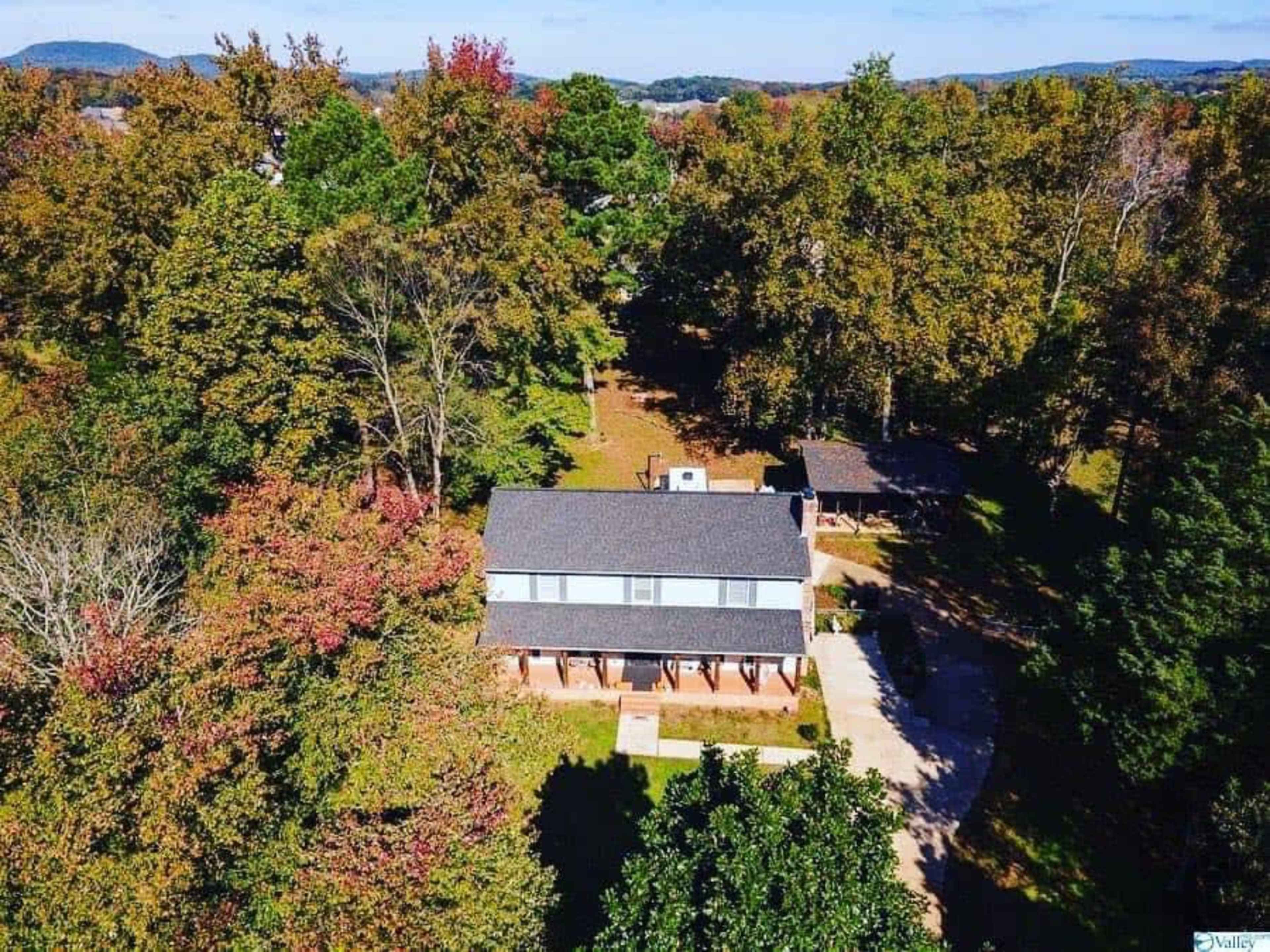 An aerial view of a house surrounded by trees with autumn foliage and a driveway leading to a secondary structure nearby.