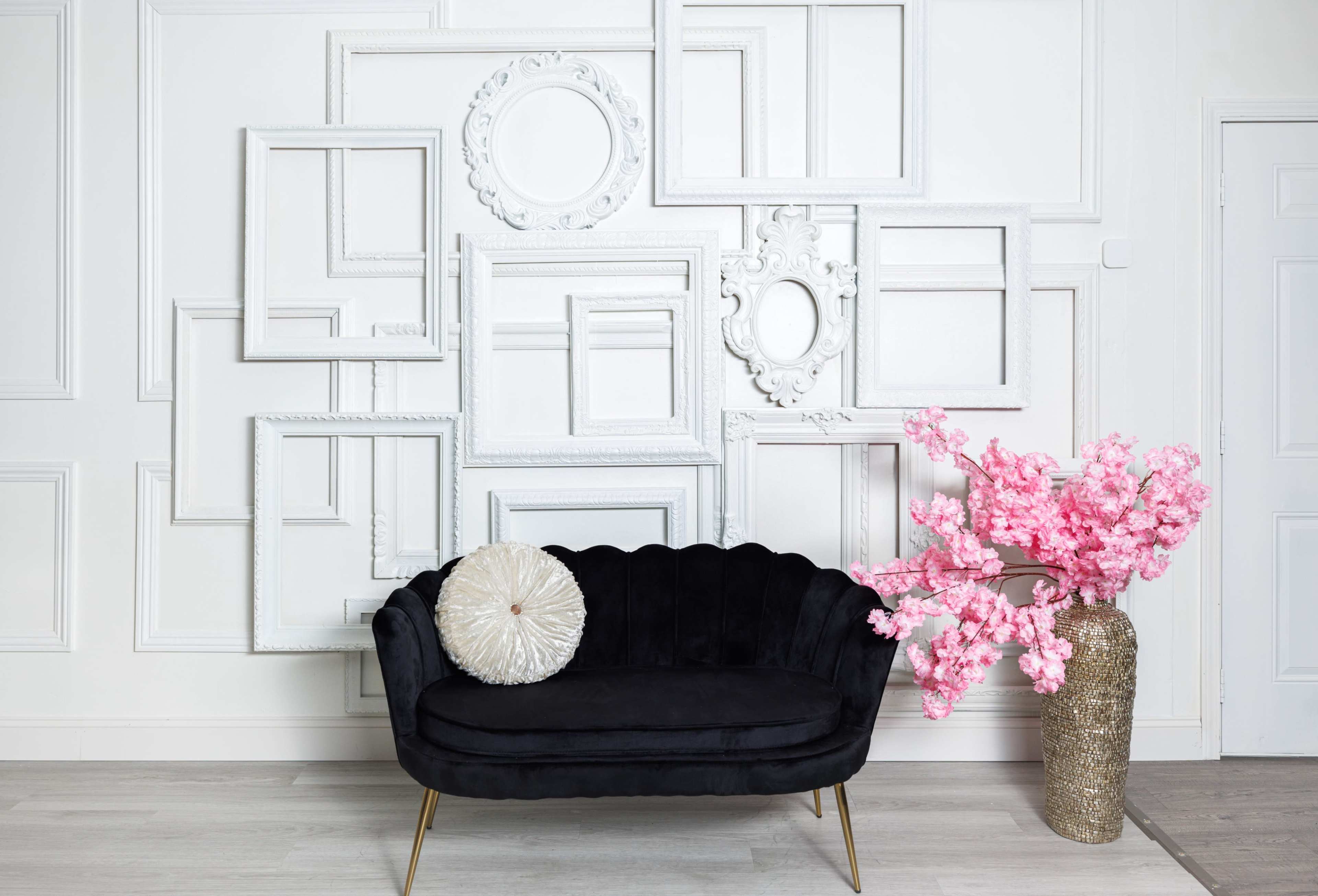 A black velvet sofa with a decorative pillow is placed in front of a wall adorned with an arrangement of white empty picture frames and a pink flower arrangement in a gold vase.