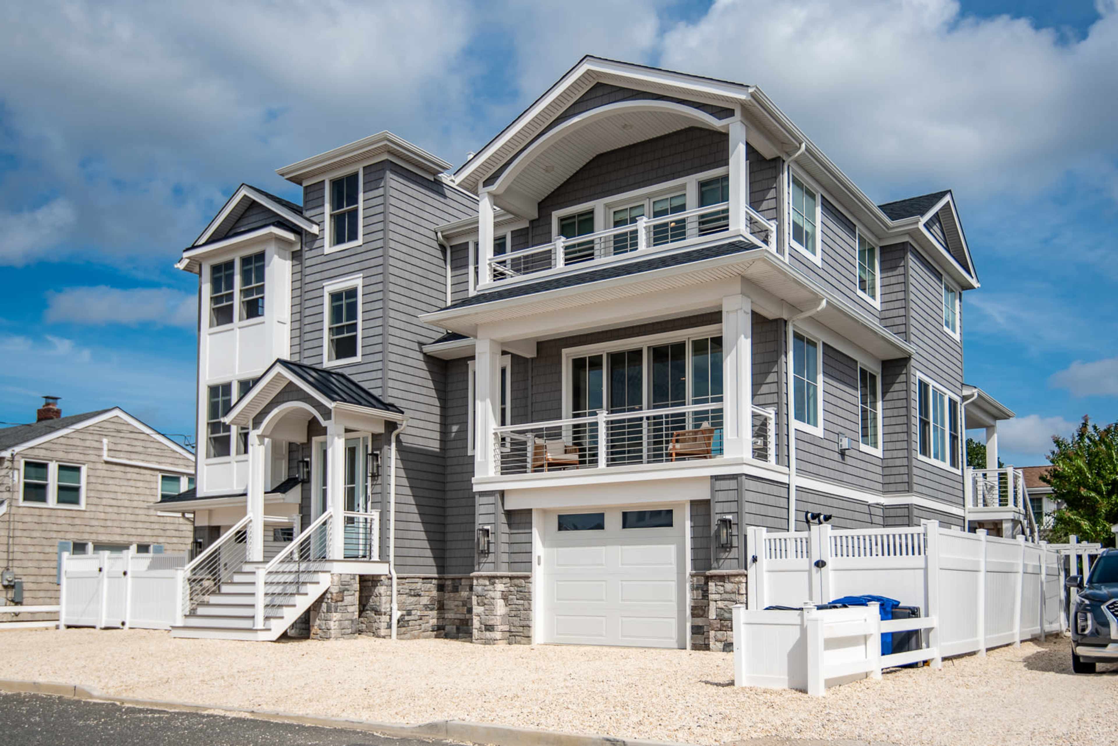 A modern, two-story coastal house with gray siding, large windows, and a spacious front porch, situated on a gravel driveway.