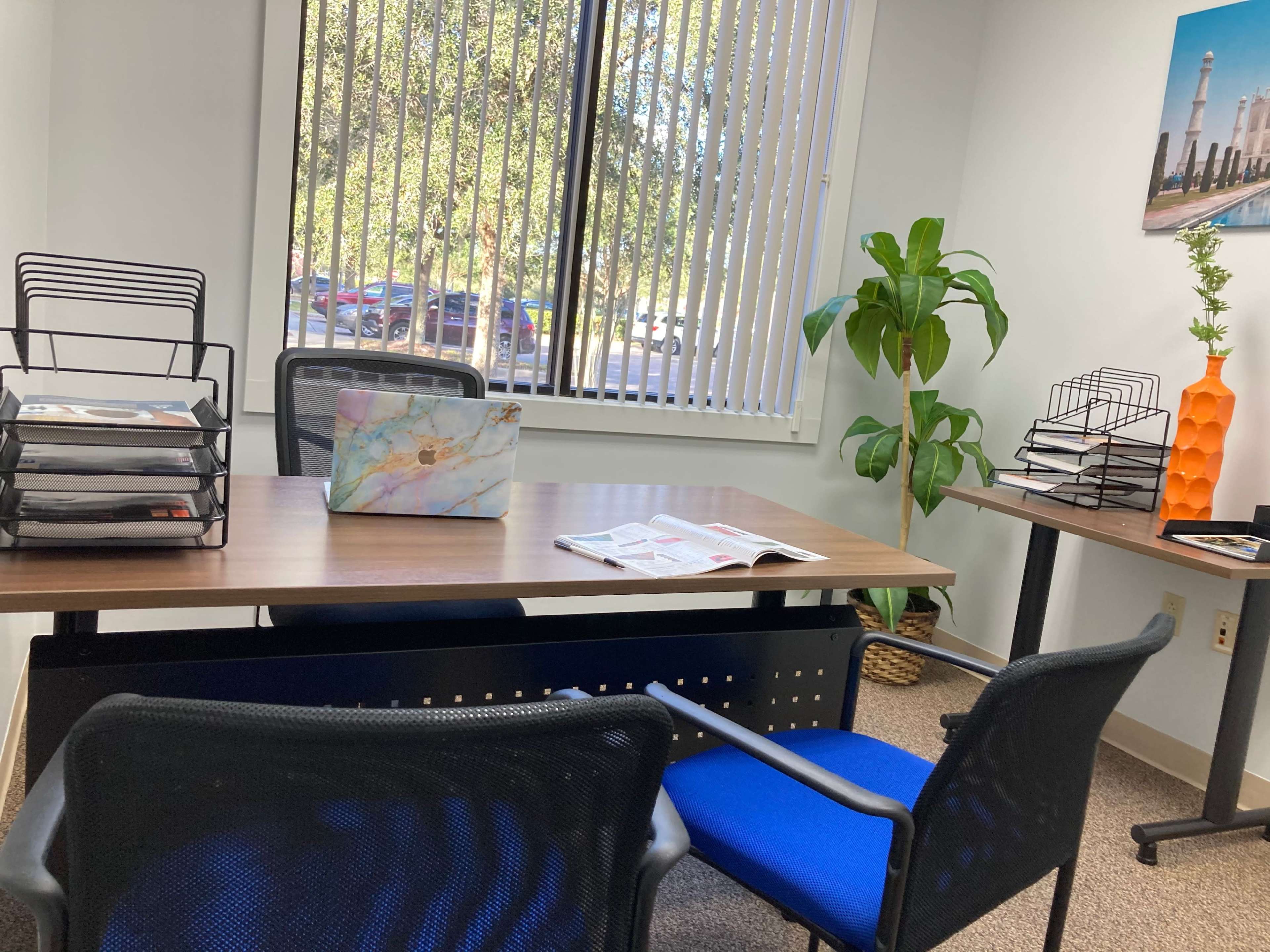 An office space featuring a wooden desk with a laptop, storage racks, and a potted plant near large windows with vertical blinds.
