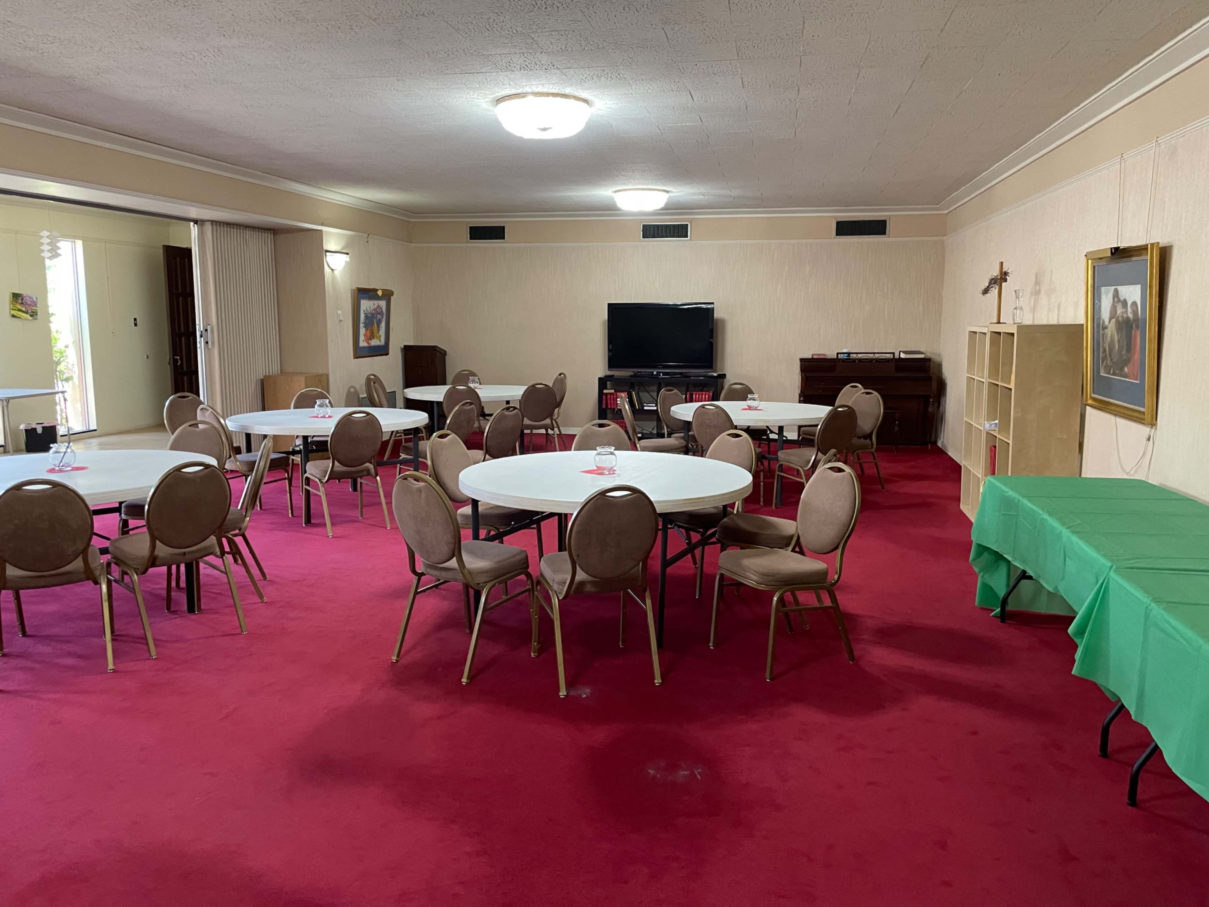 A spacious room with a red carpet, featuring several round tables surrounded by chairs, a television, a piano, and a green table along one wall.