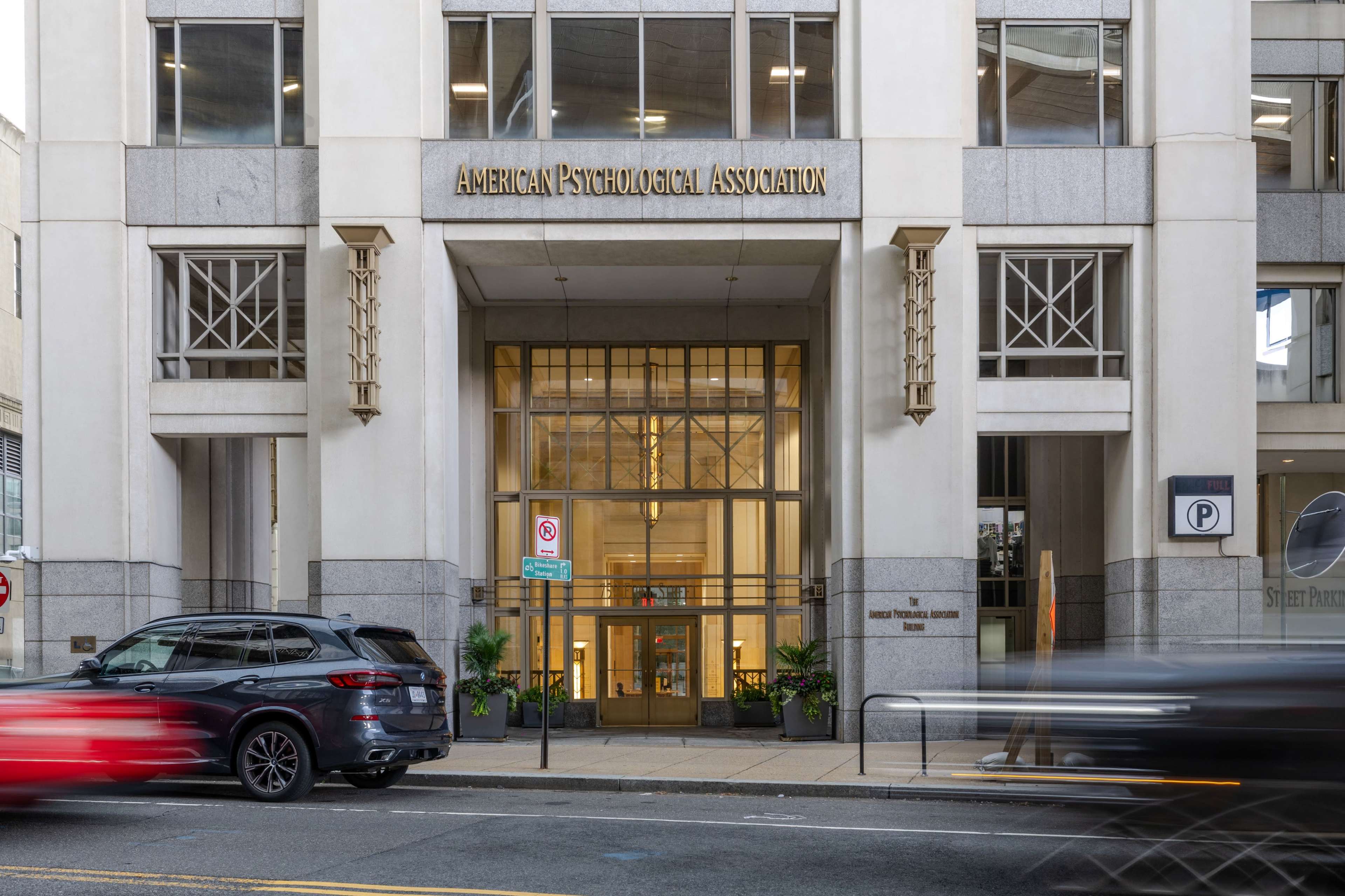 The image shows the exterior of the American Psychological Association building, featuring a modern entrance with large glass doors and the organization's name displayed prominently above.