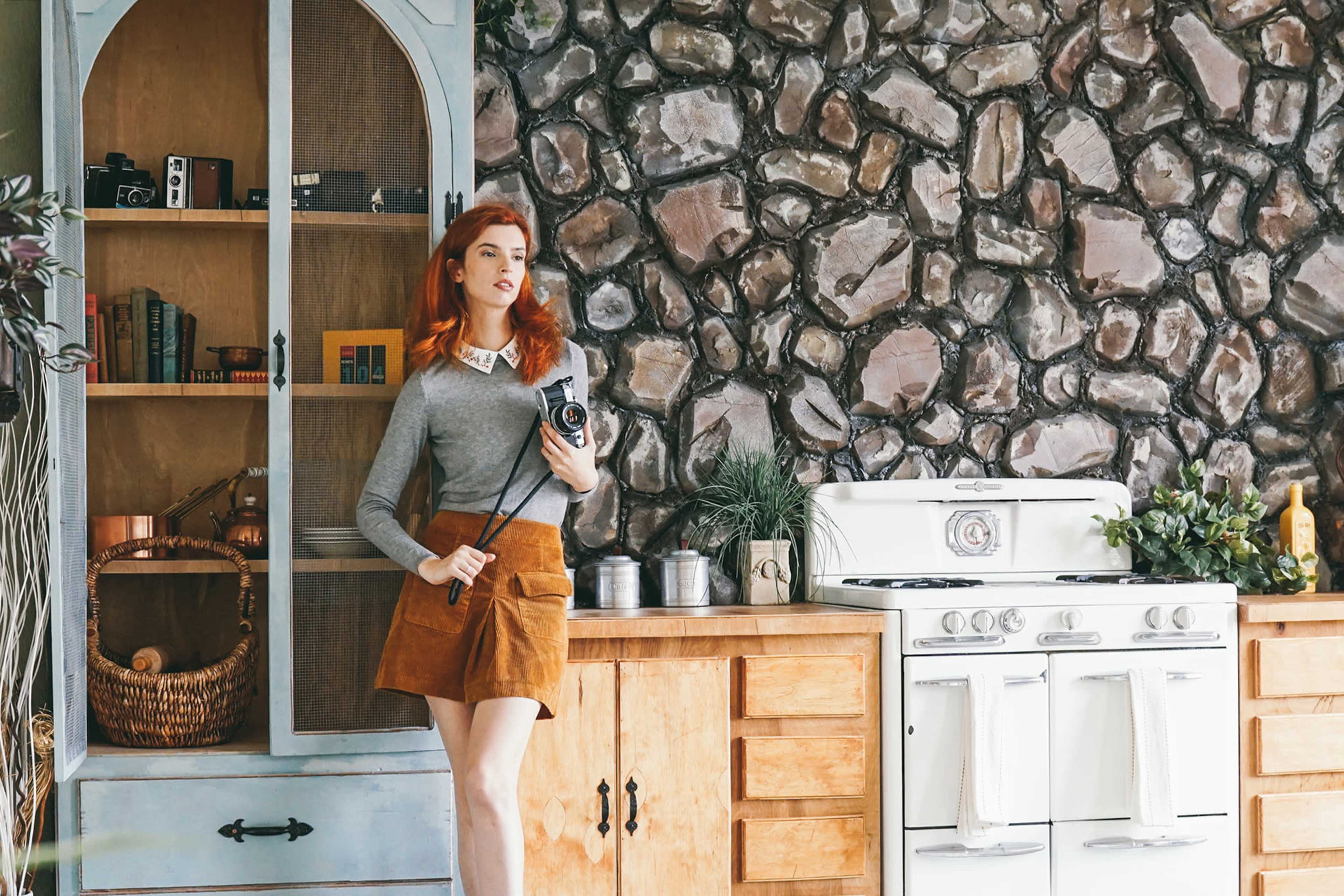A woman with red hair stands in a vintage kitchen with a stone wall, holding a camera and wearing a gray sweater and brown skirt.