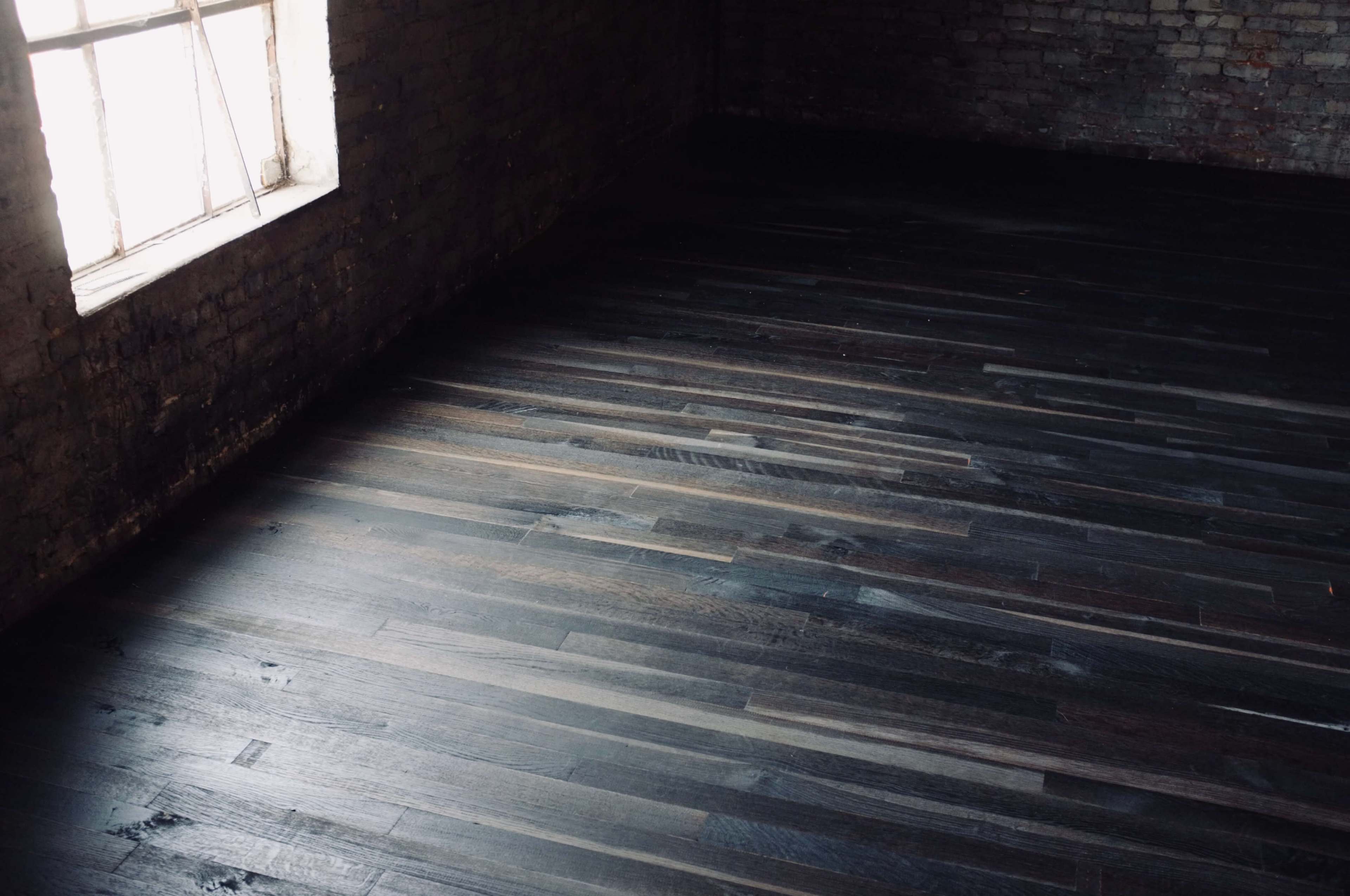 The image shows a dimly lit interior space with dark wooden flooring and a single window allowing soft light to enter.
