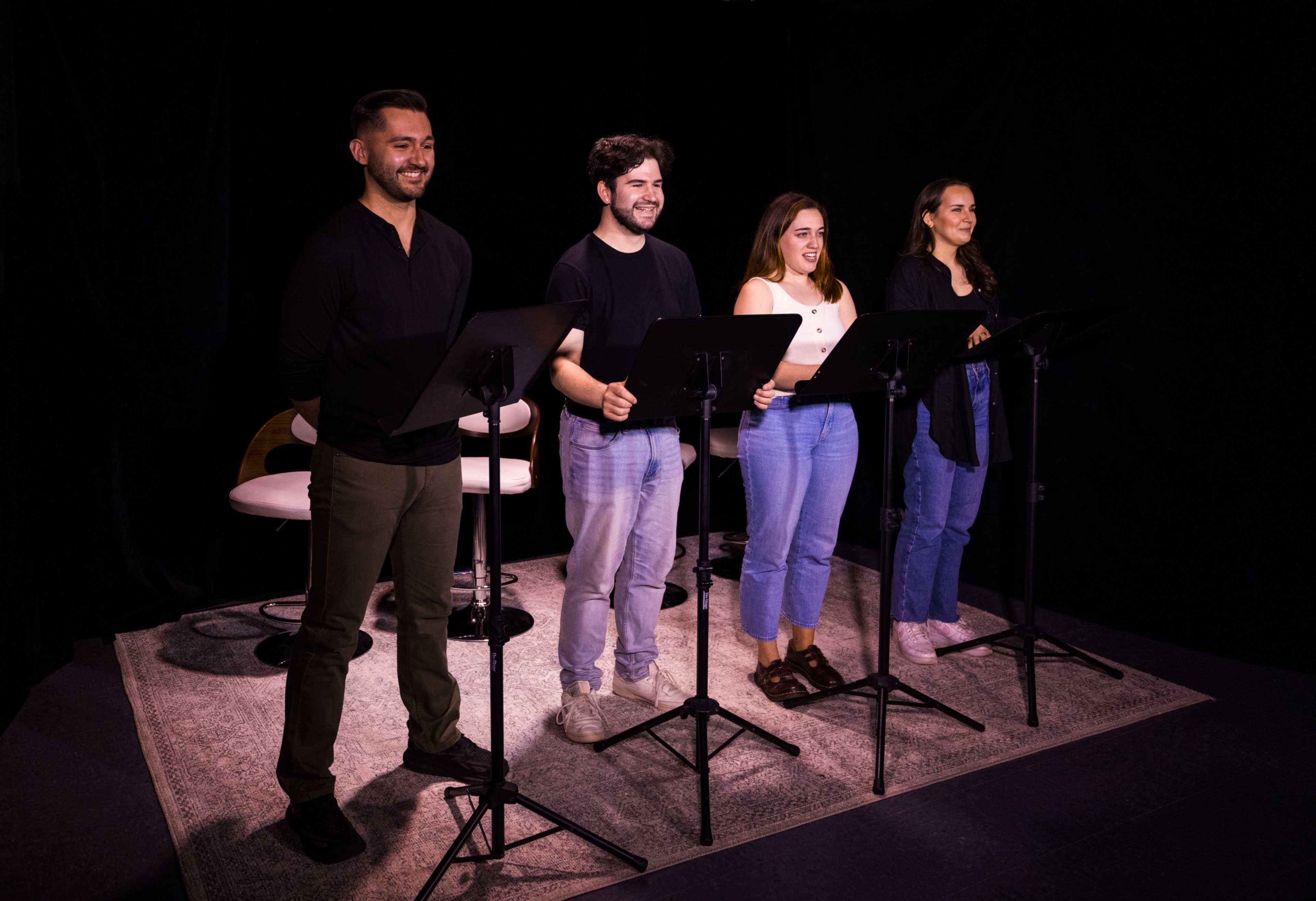 Four performers stand behind music stands on a stage, reading from scripts under soft lighting.