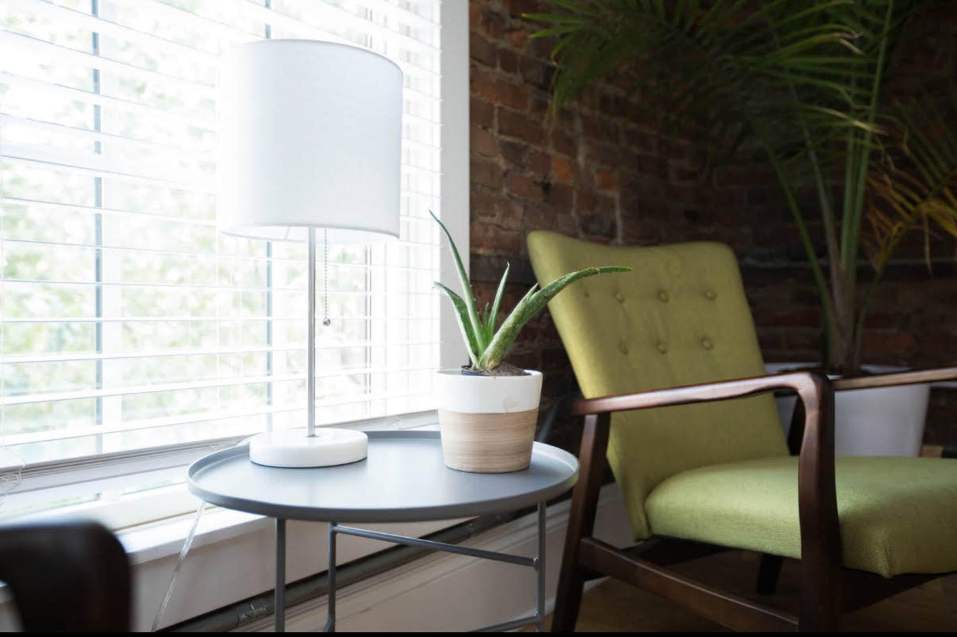 A table lamp and a potted plant sit on a side table next to a green armchair by a window with blinds.