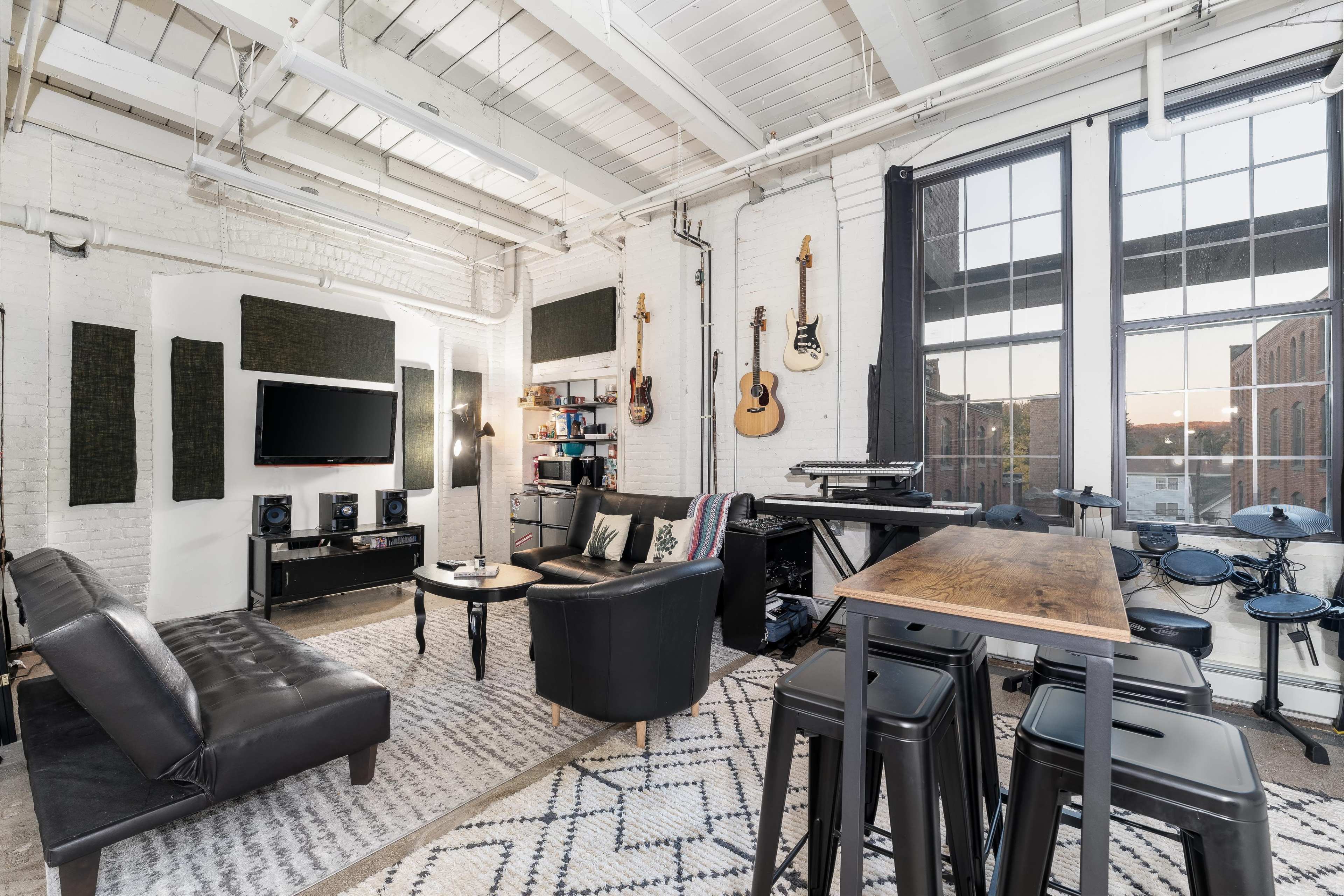 A modern living space with a black leather sofa, a wall-mounted TV, musical instruments hanging on the walls, and a wooden table with high stools.