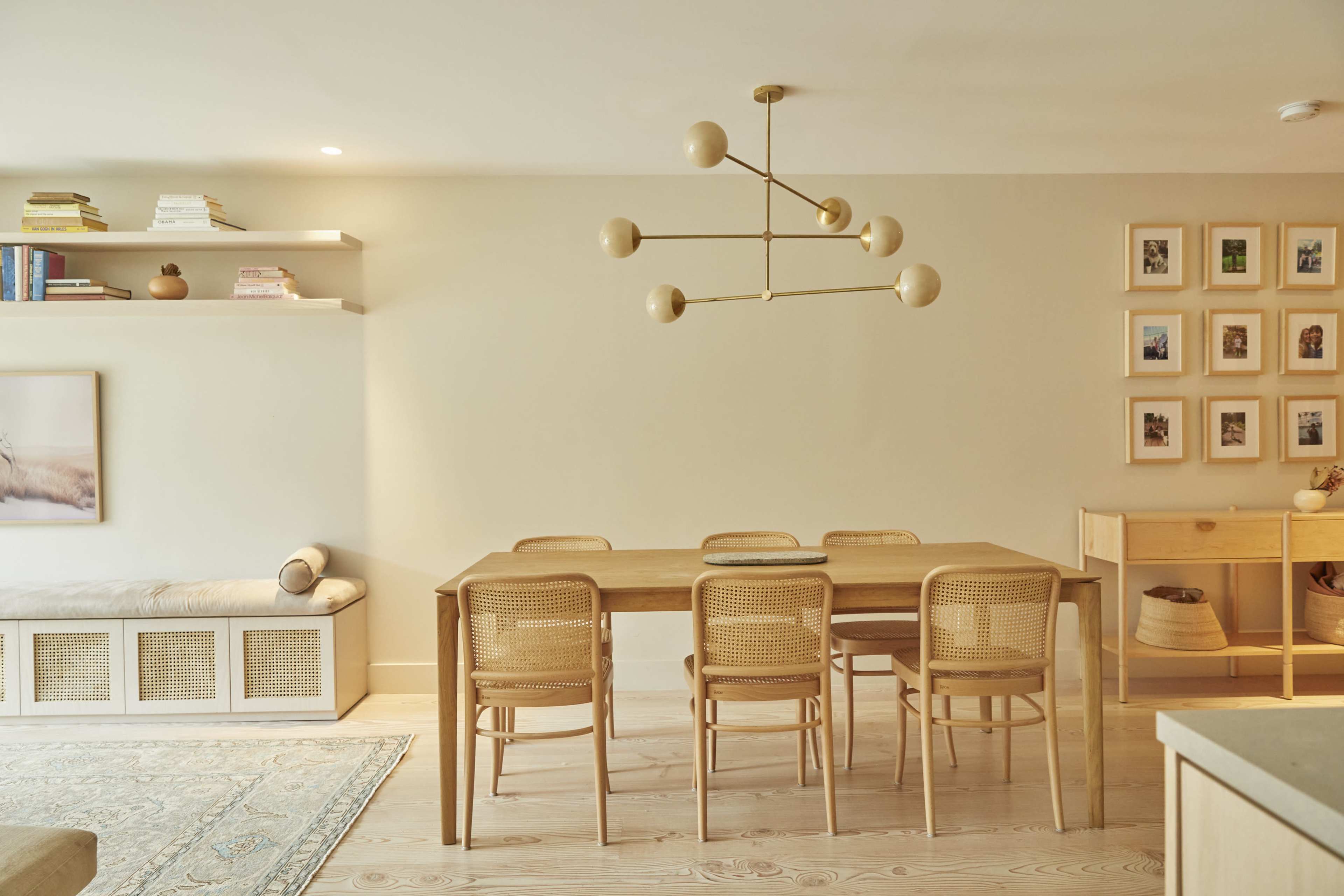 A modern dining area features a wooden table surrounded by six chairs, a light fixture with multiple bulbs overhead, and shelves with decorative items and framed photos on the walls.