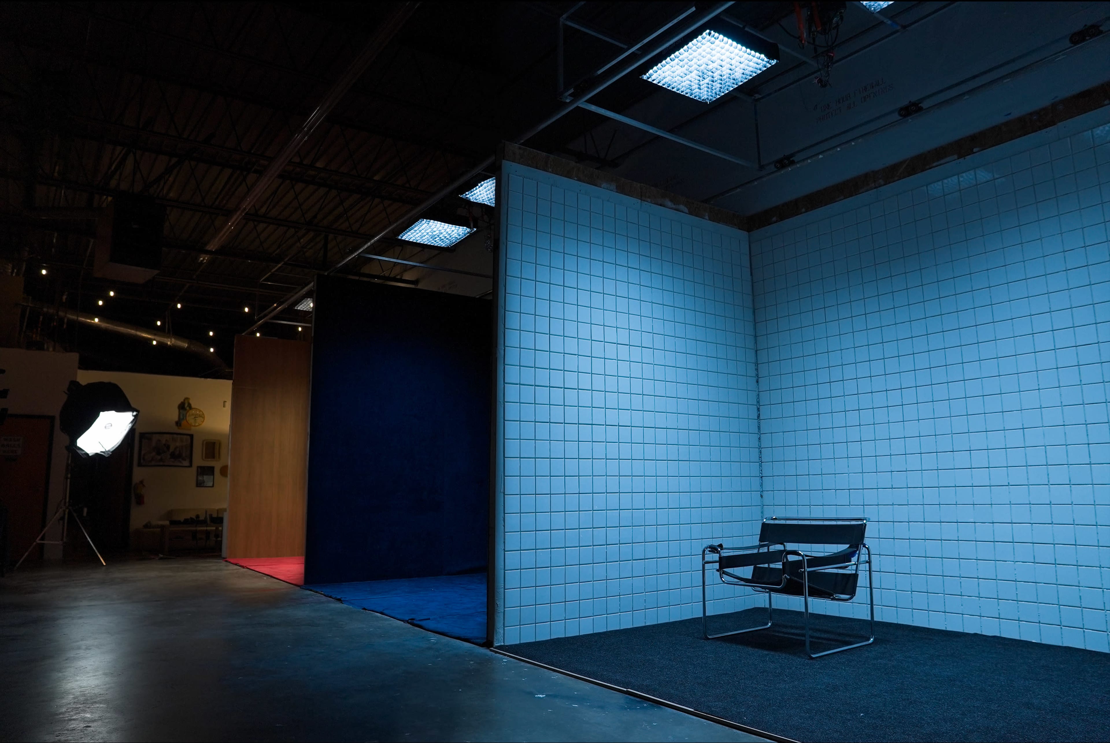 The image shows a minimalist studio space featuring a single chair positioned on a dark carpet, surrounded by walls of white and colorful panels under various lighting.