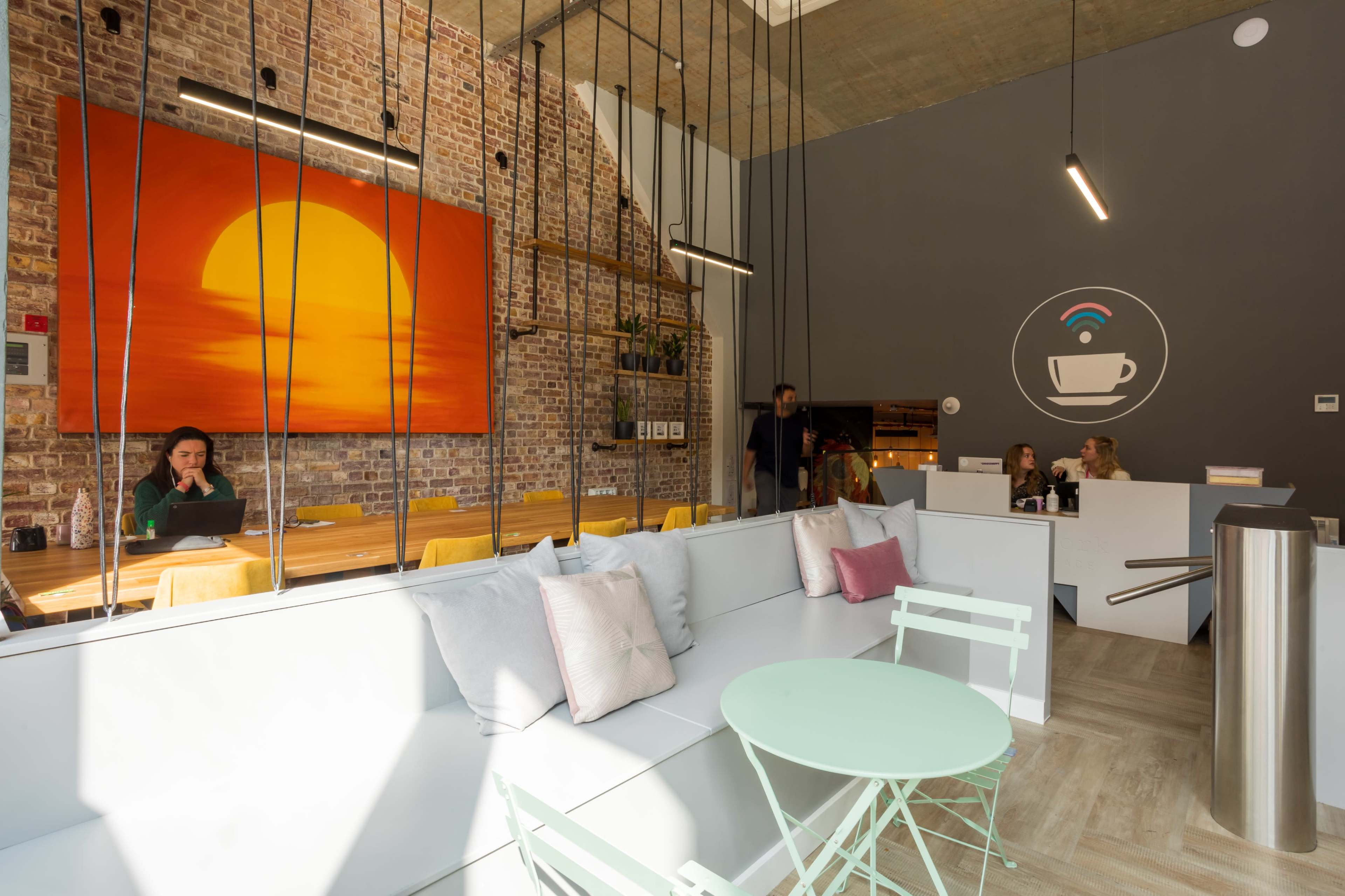 A modern café features a large orange sunset painting on a brick wall, with stylish seating and patrons engaged in conversation.