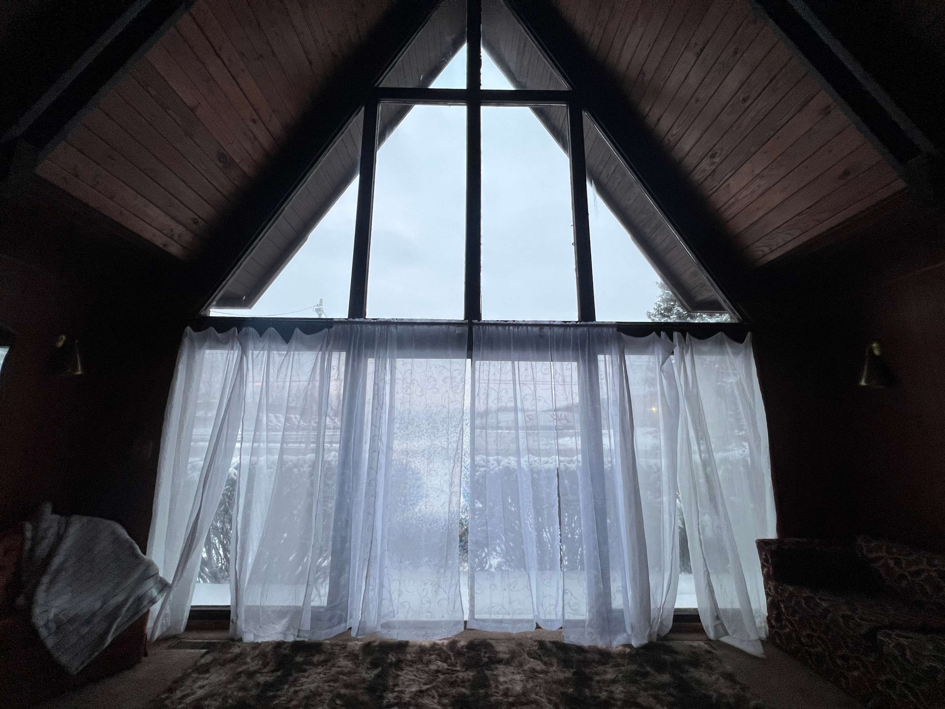 A large A-frame window with sheer curtains overlooks a wintery landscape.