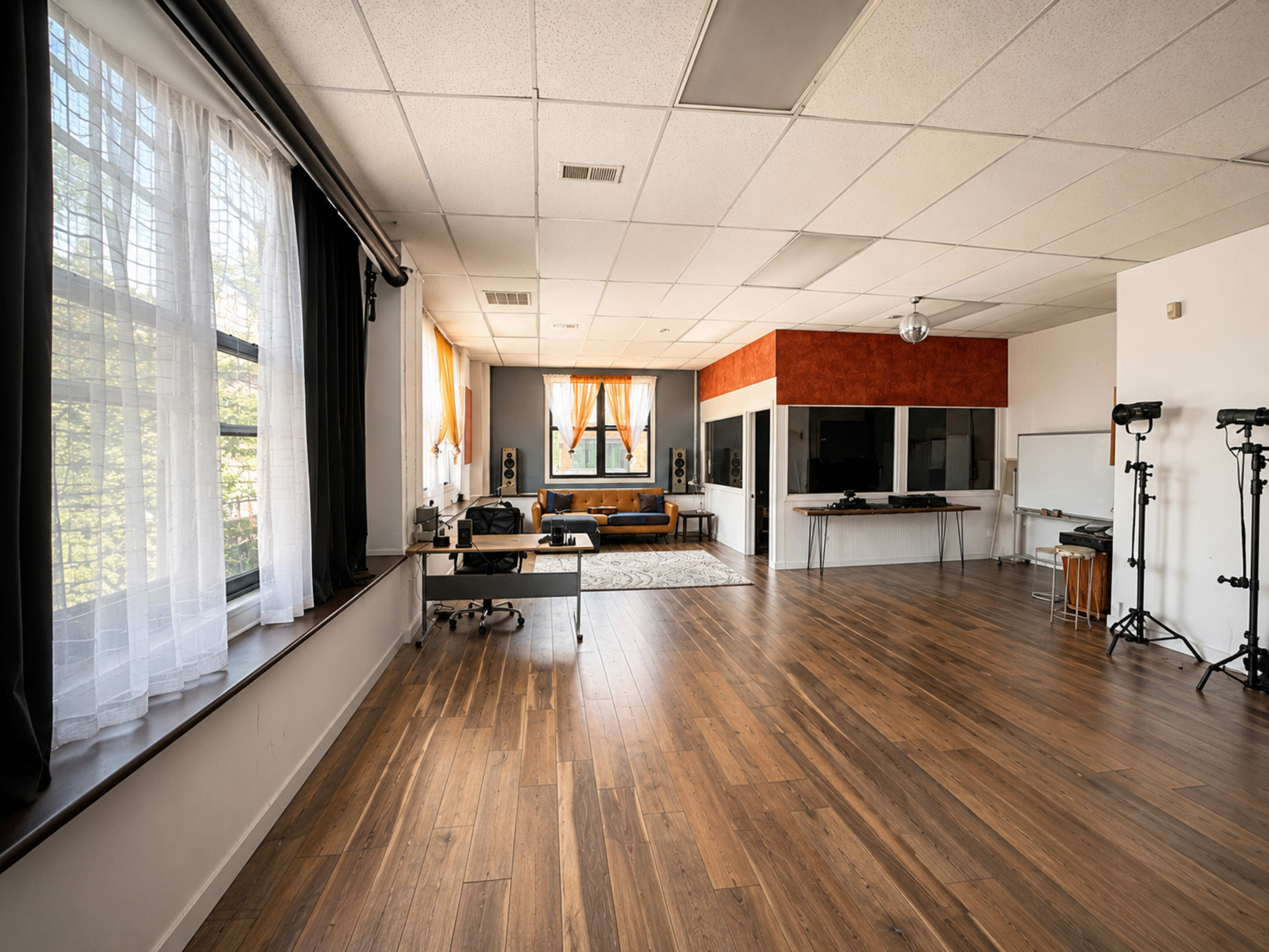 The image shows a spacious, well-lit interior room with wooden floors, large windows, and a mix of furniture, including a couch and music equipment.