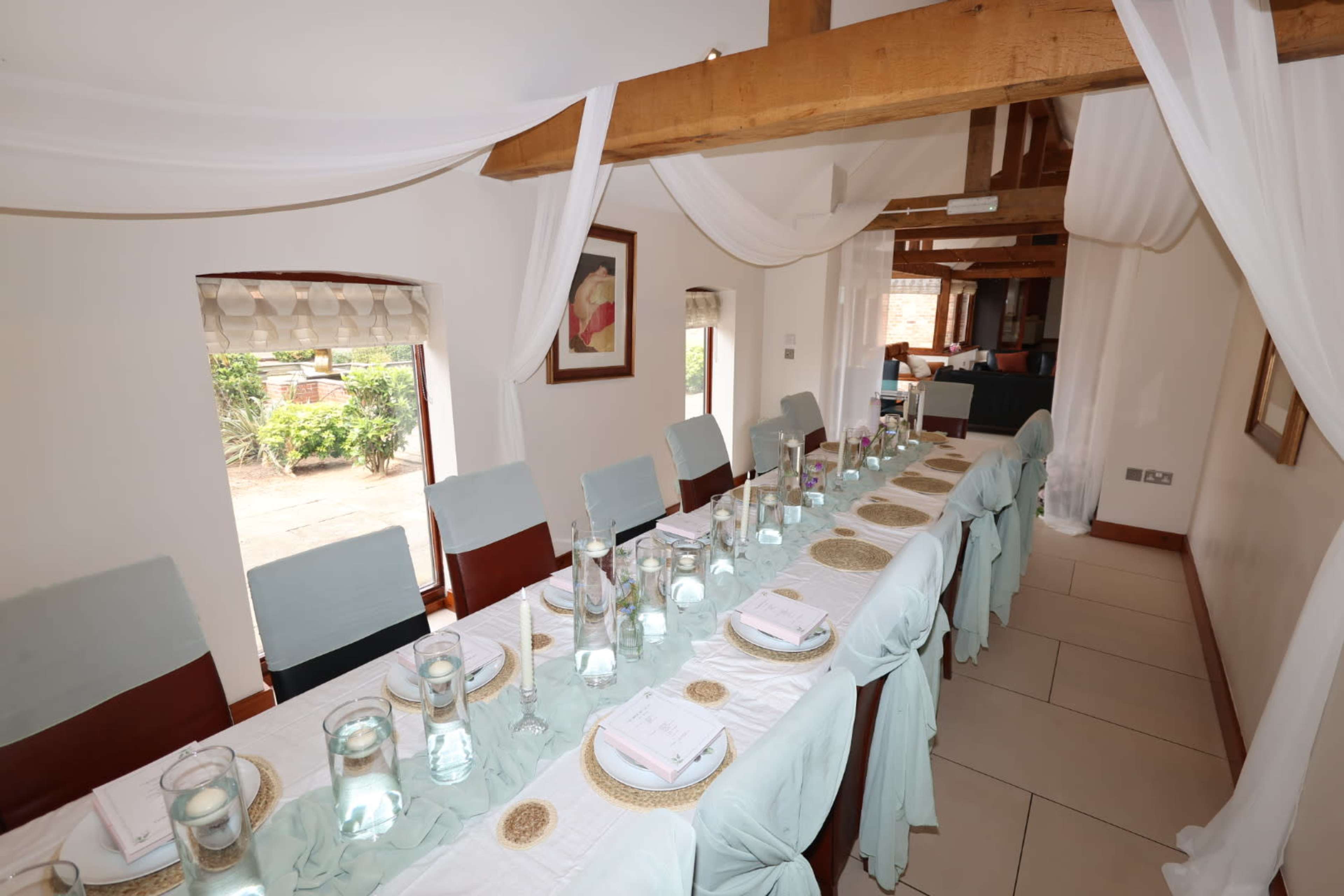A long dining table is set formally with clear water pitchers and glasses, surrounded by chairs, and draped with light fabric in a bright, open space.