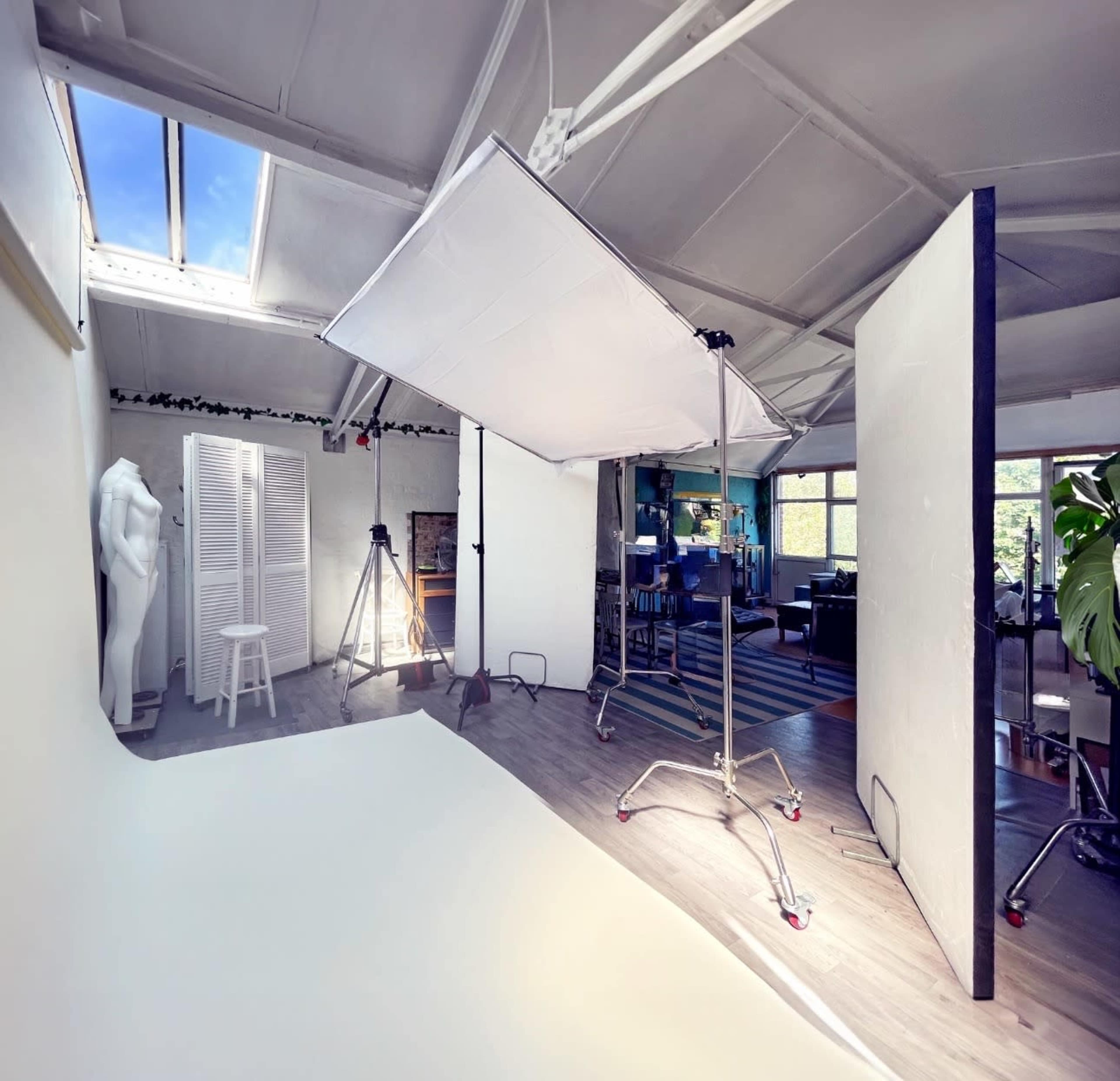 The image shows a photography studio with a large softbox, a white backdrop, and various equipment set up in a spacious room featuring large windows and minimal furnishings.
