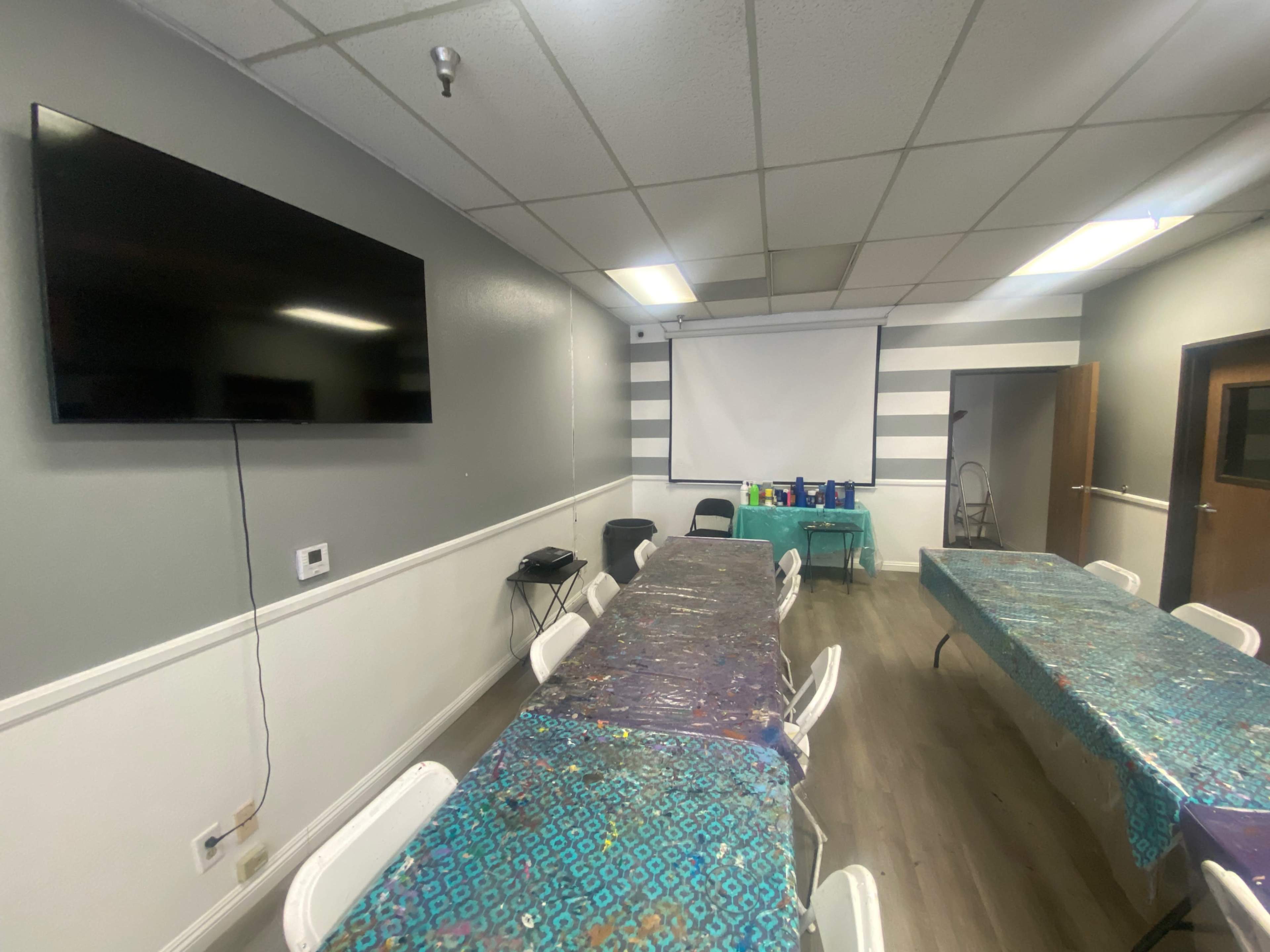 A room equipped with several tables covered in blue tablecloths, a large wall-mounted television, and a pull-down projector screen.