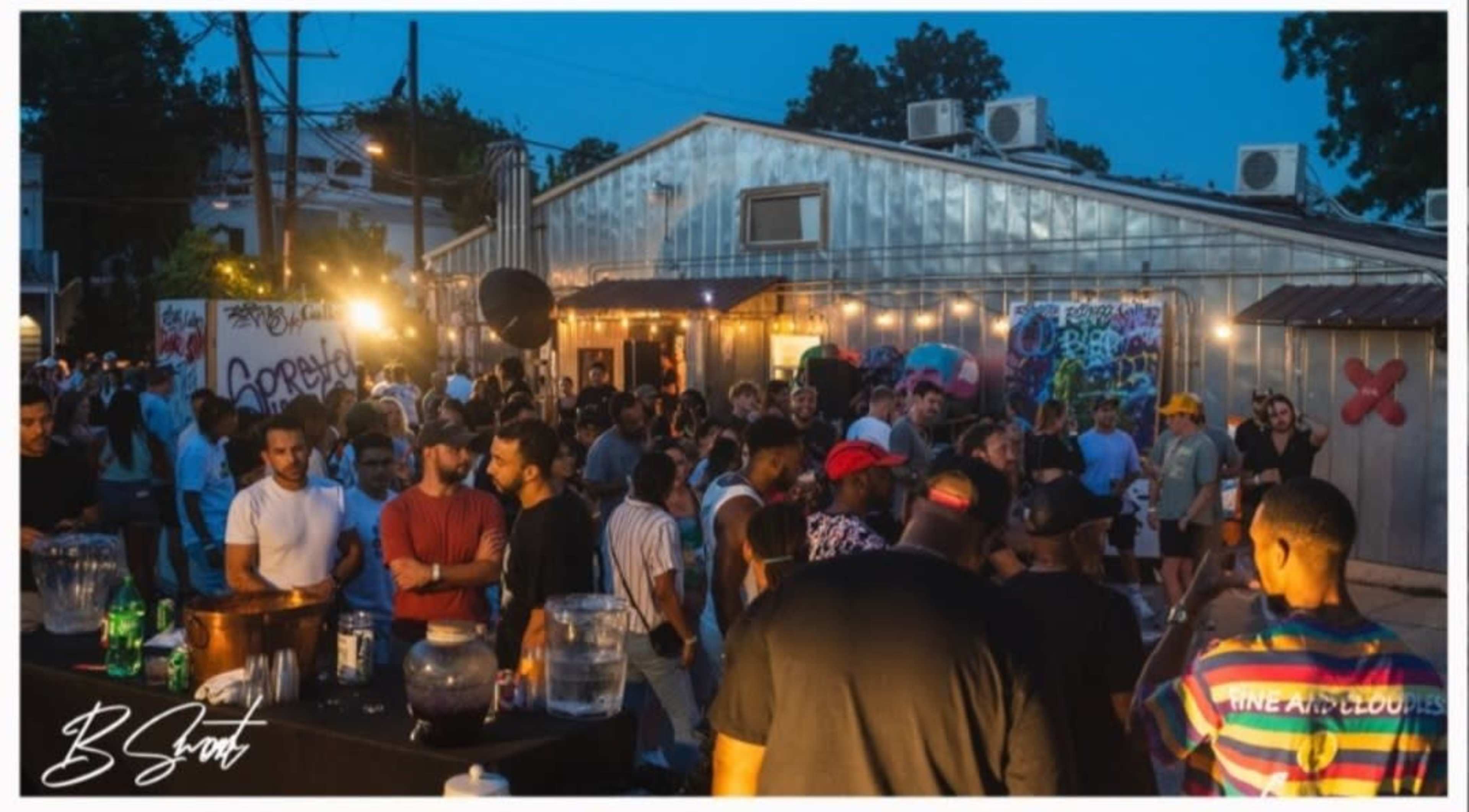 A large group of people socializes outside a venue with lights, food, and drinks during an evening event.