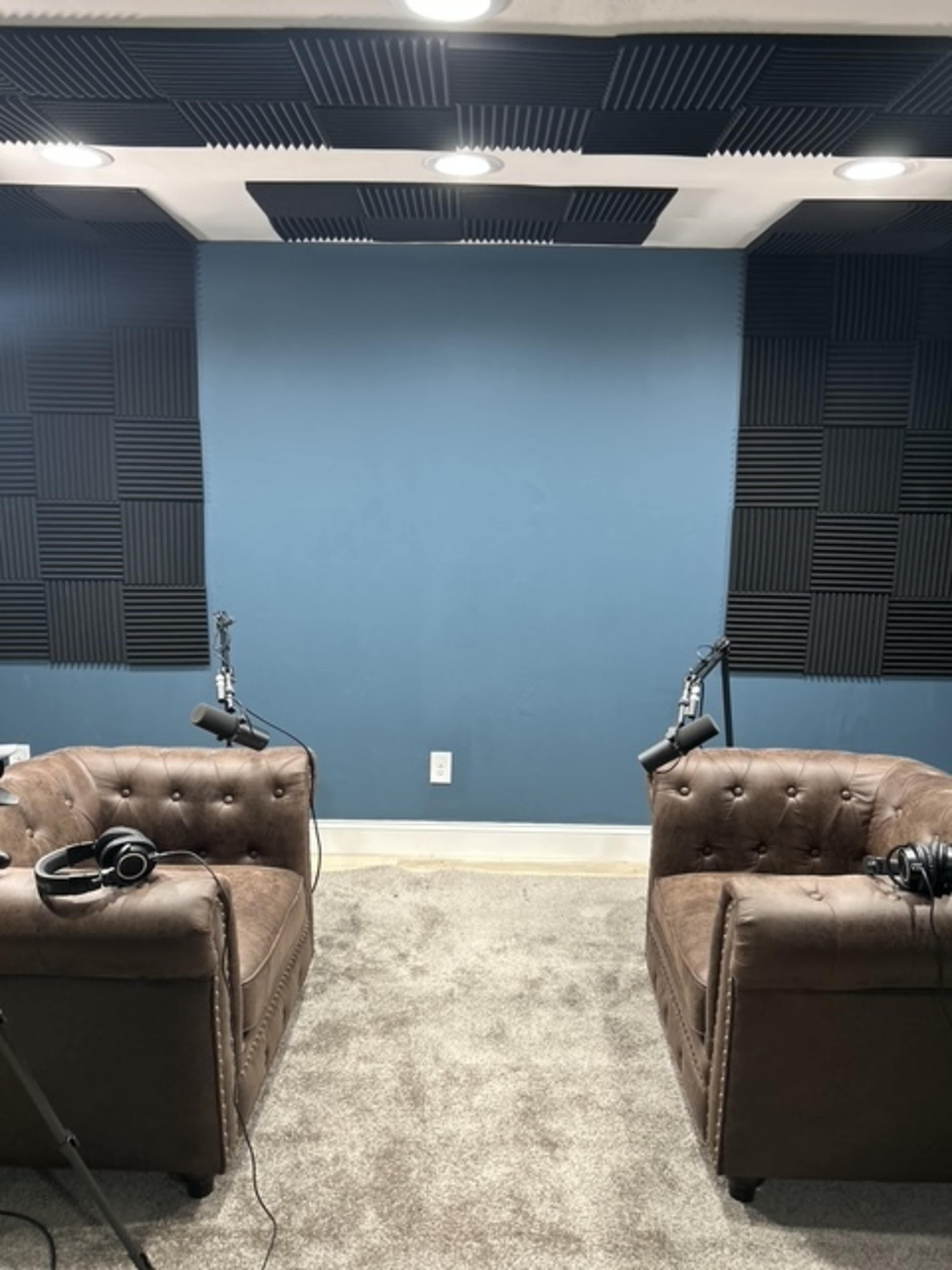 The image shows a small recording studio with two brown upholstered chairs, each equipped with a microphone and headphones, facing a blue wall.