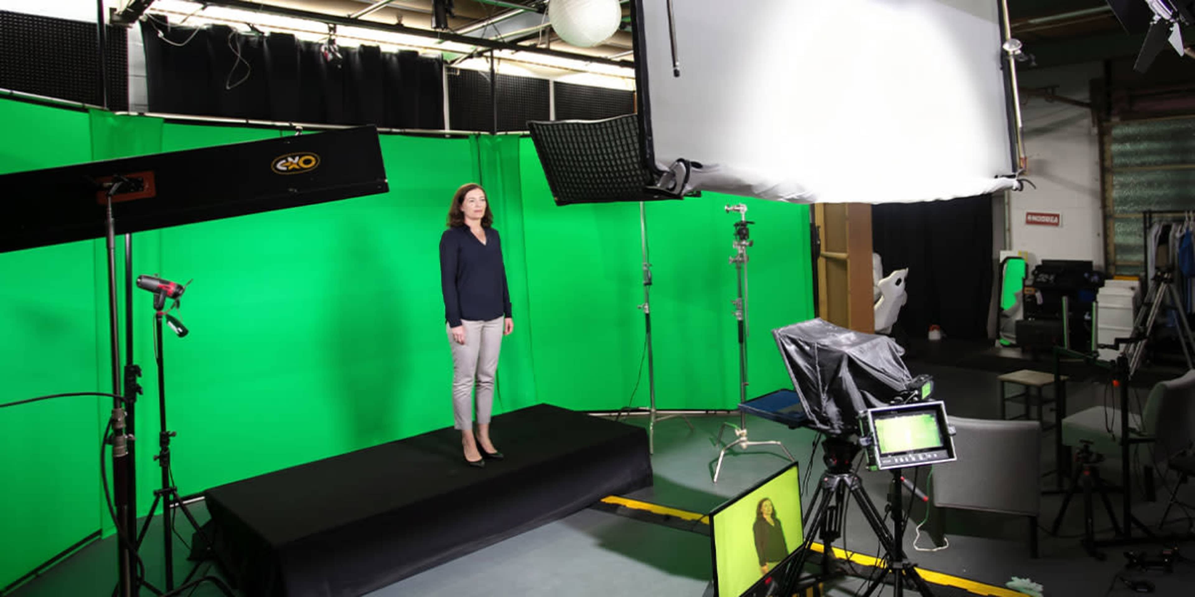 A female presenter stands on a platform in front of a green screen in a studio setting, with multiple lighting and camera equipment arranged around her.