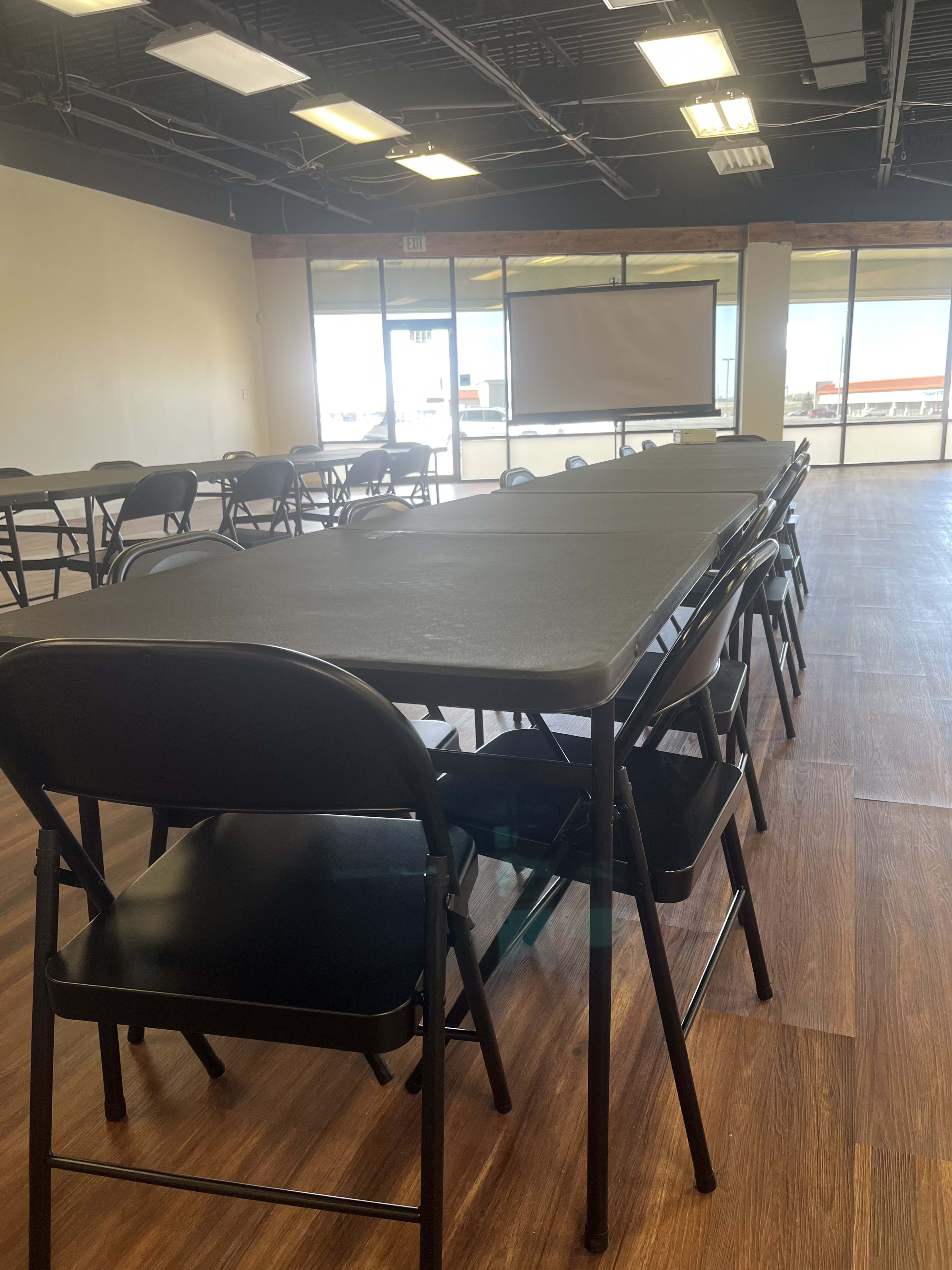 A spacious room with multiple rows of black folding tables and chairs, facing a large projector screen.