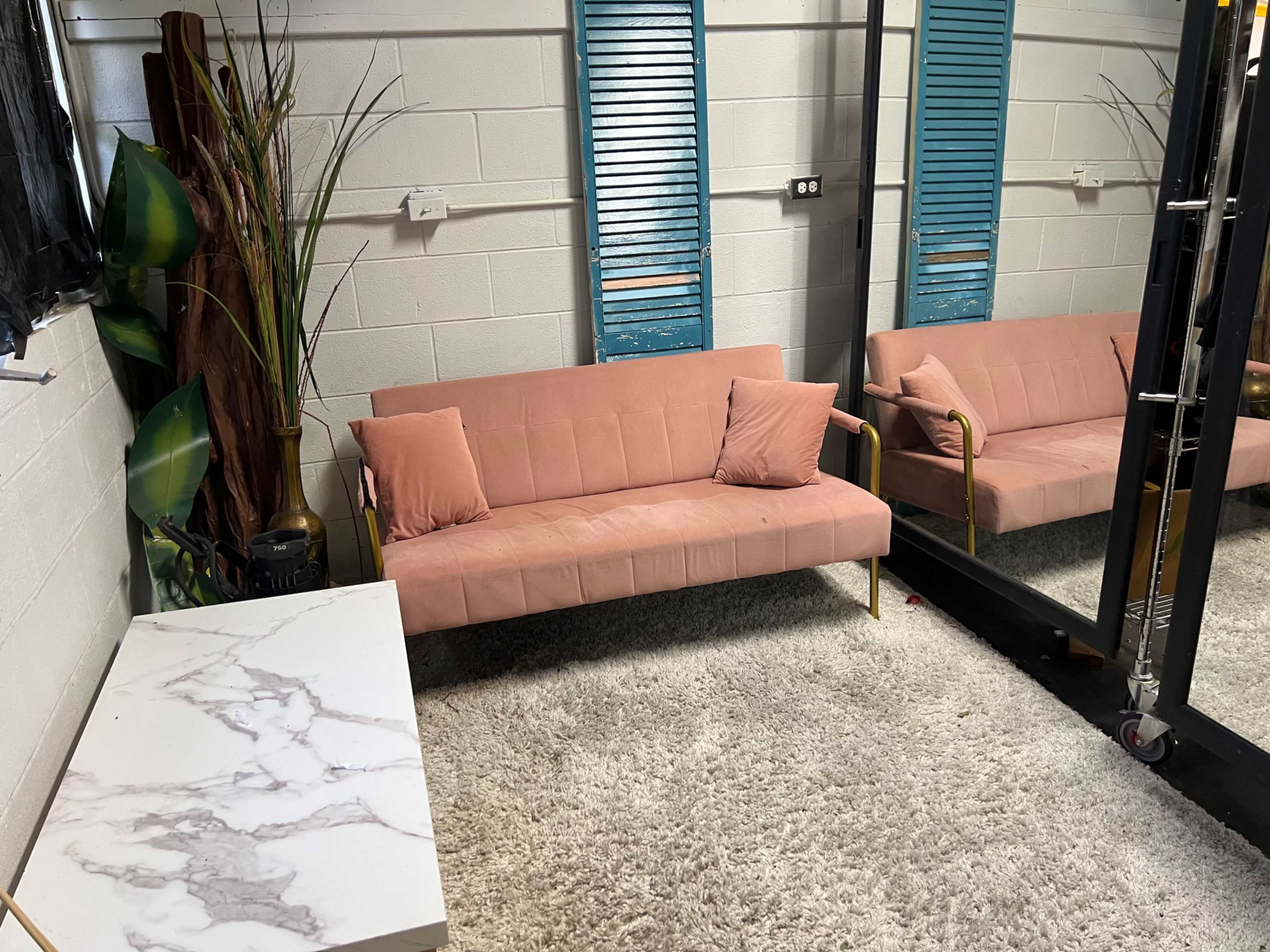 The image shows a cozy room with a pink sofa, a marble-patterned table, and a large mirror against the wall, featuring decorative plants and blue shutters.