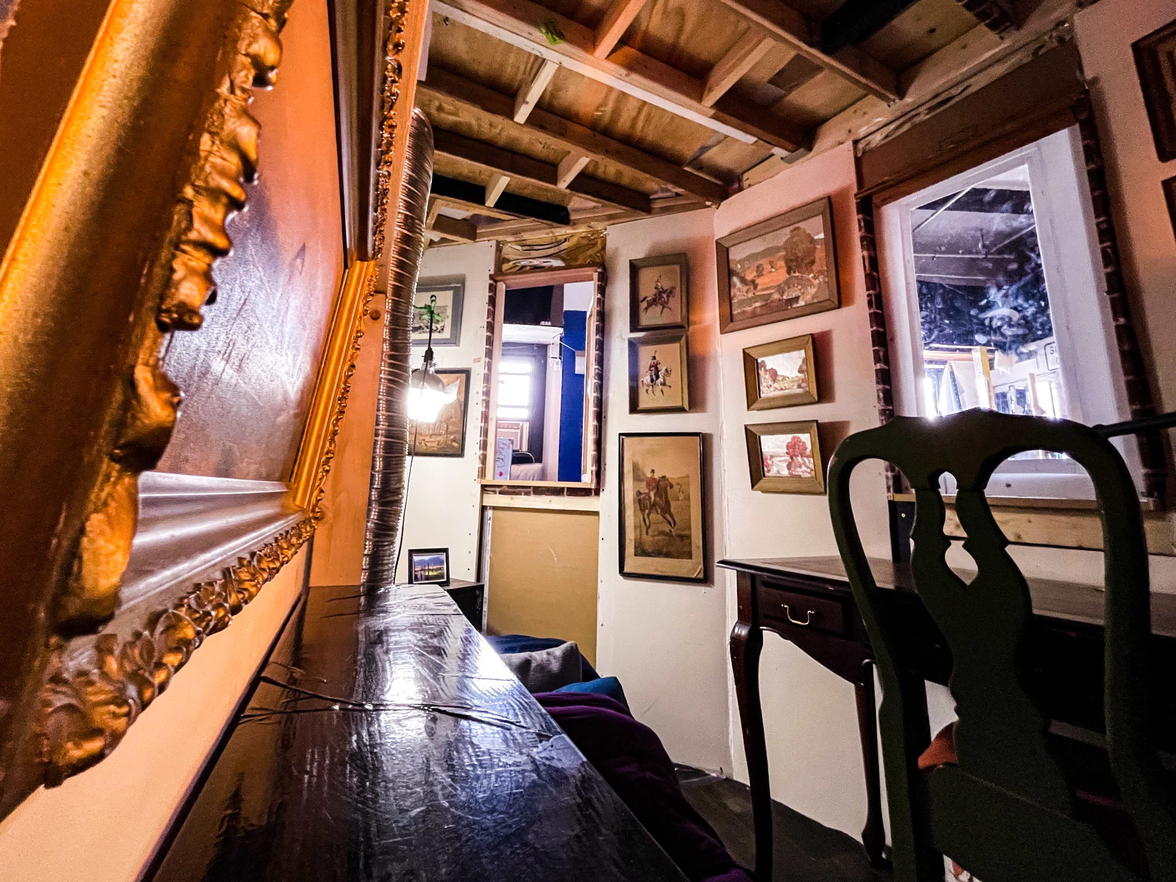 The image shows a cozy interior of a room with wooden beams, framed artwork on the walls, and a dark table with a green chair.
