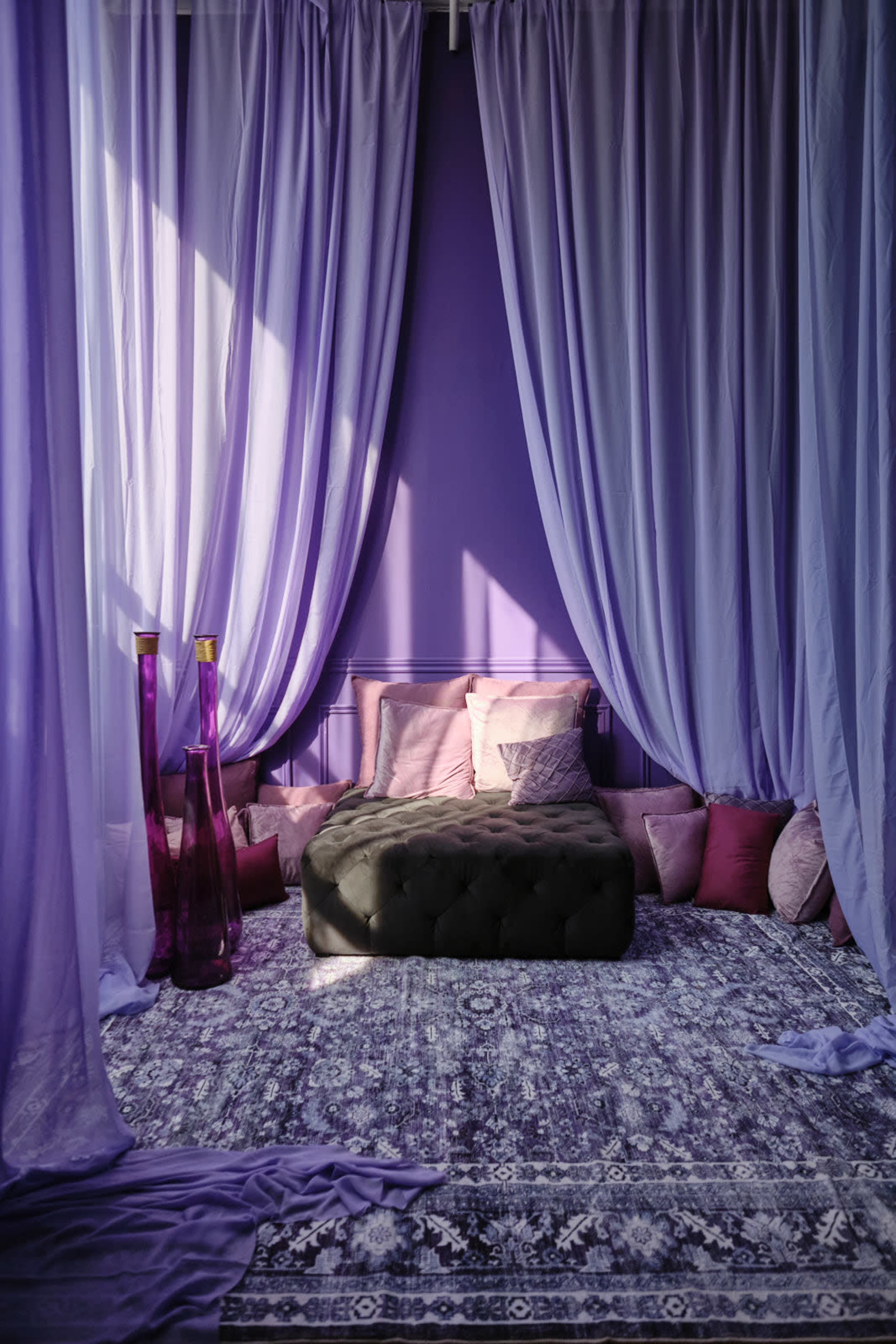 The image shows a cozy corner of a room with purple curtains, a tufted seating area, and various cushions on a patterned rug.