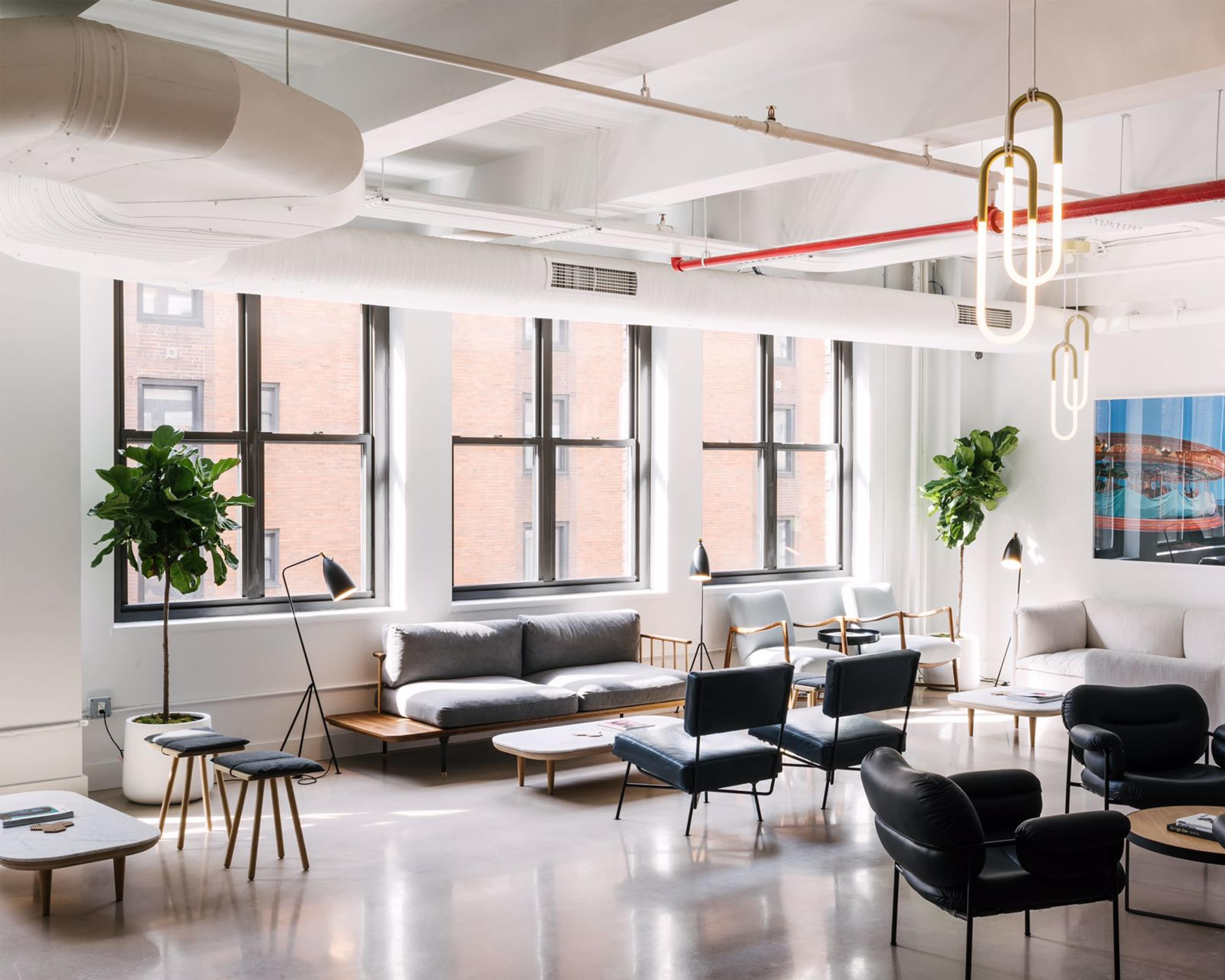 A modern lounge area features various seating options, potted plants, and multiple windows allowing natural light to illuminate the space.