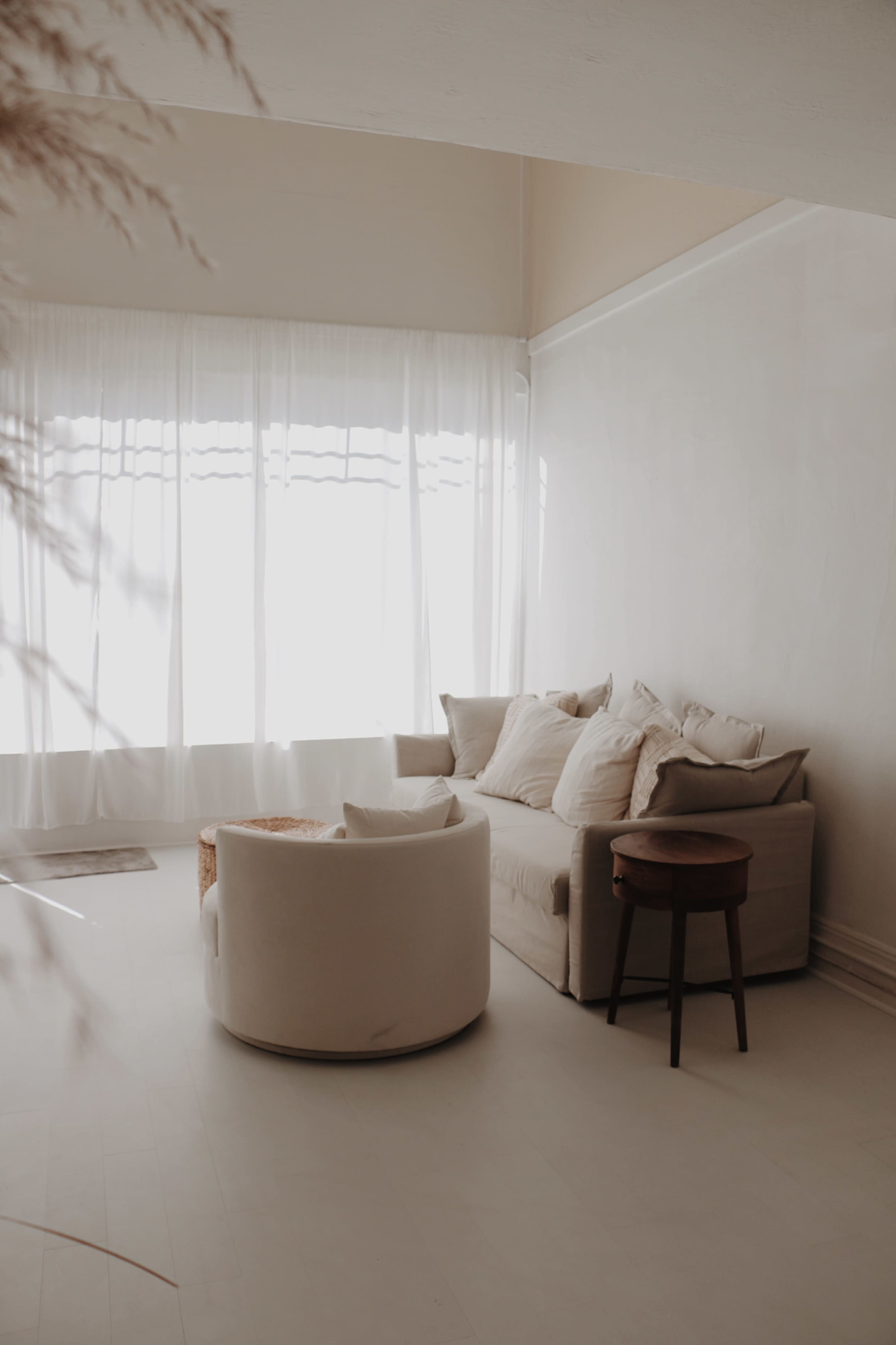 A minimalist living room features a light-colored sofa with cushions, a round chair, and a small wooden side table near a large window with flowing curtains.